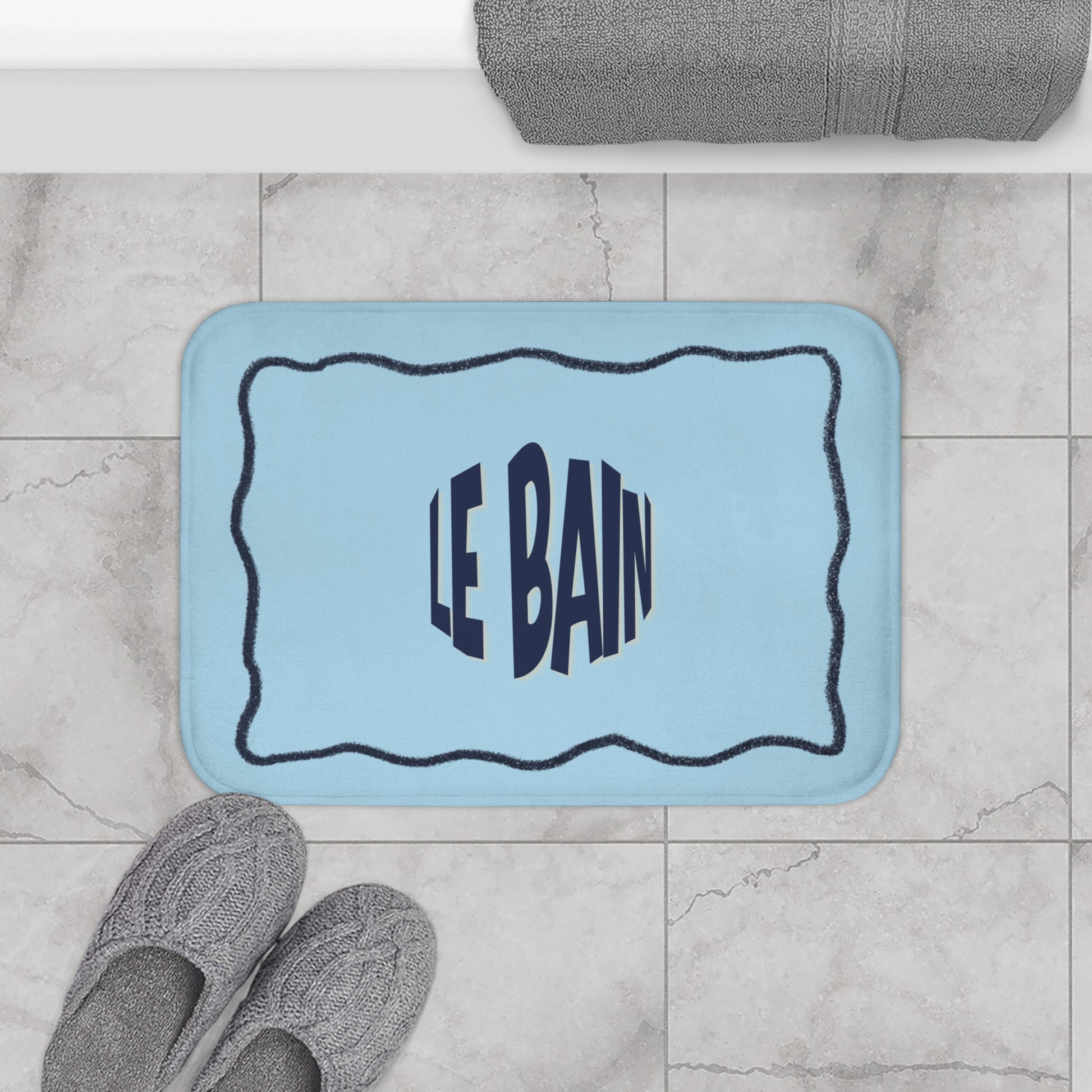 Light blue bath mat with 'LE BAIN' text on a tiled bathroom floor.