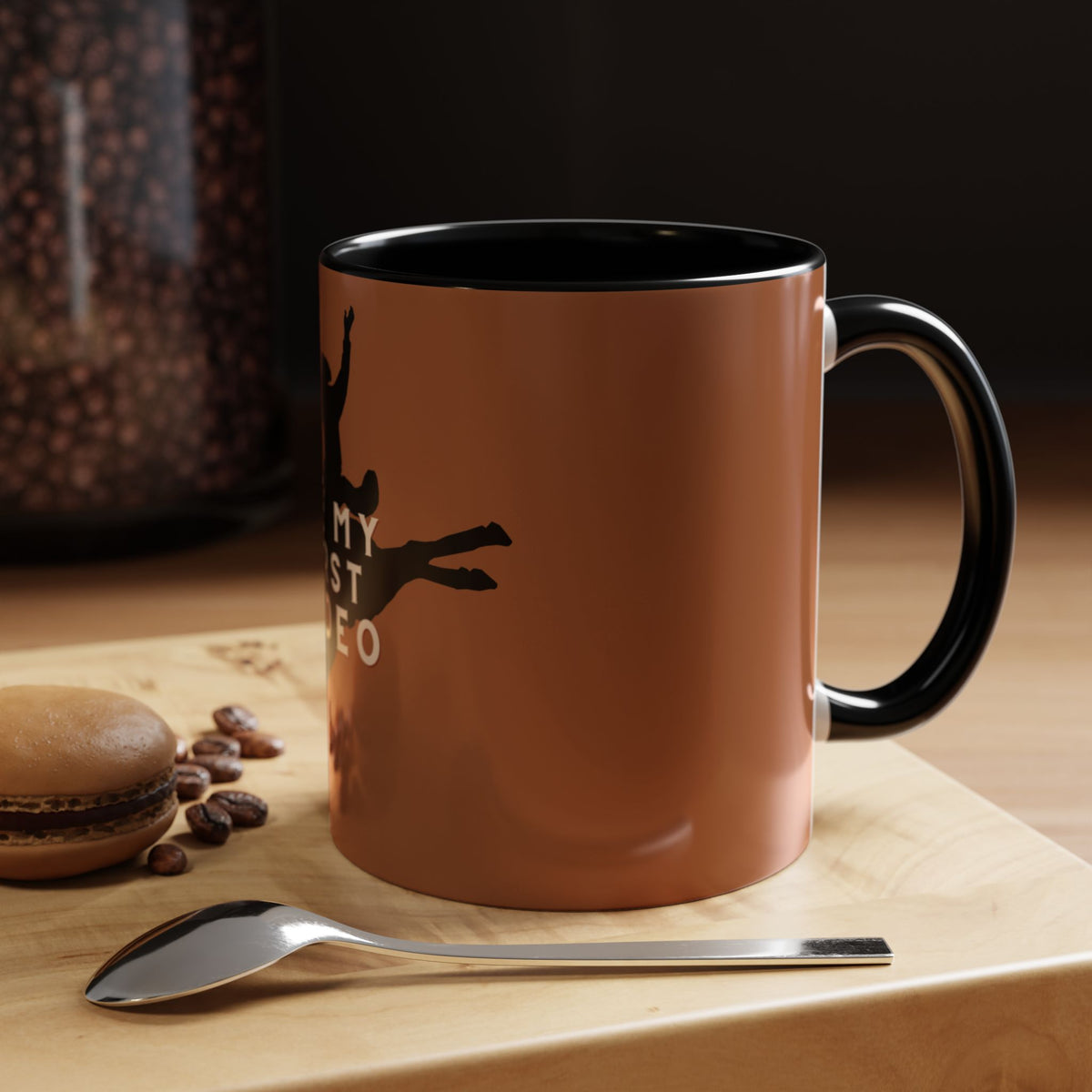 A ceramic coffee mug with a glossy terracotta brown exterior, black interior, and black handle, featuring a black silhouette of a person jumping with the text 'MY FIRST JUMBO'. The mug is set on a wooden board next to a spoon, coffee beans, and a macaron, with a dark background.