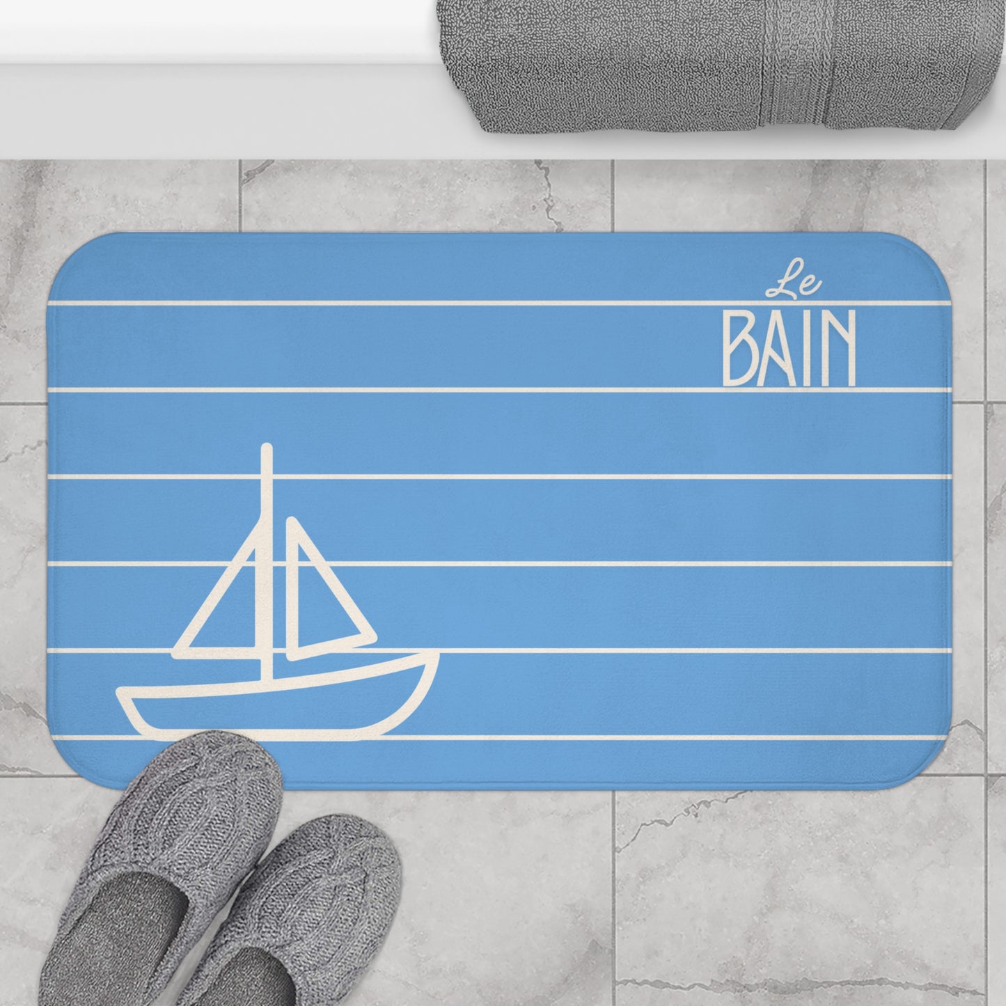 Blue bath mat with sailboat design and 'Le Bain' text on a bathroom floor.