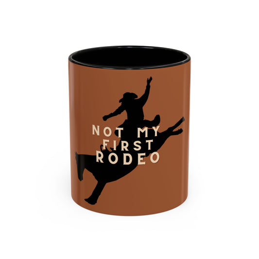 A ceramic mug with a terracotta brown exterior, black interior, and a black silhouette of a cowboy riding a bucking bull, with the text 'NOT MY FIRST RODEO' in beige.