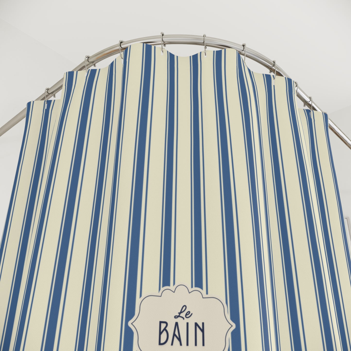 A close-up of a striped fabric shower curtain with vertical blue and cream stripes, hanging from a curved metal rod. A decorative label at the bottom reads 'Le Bain'.