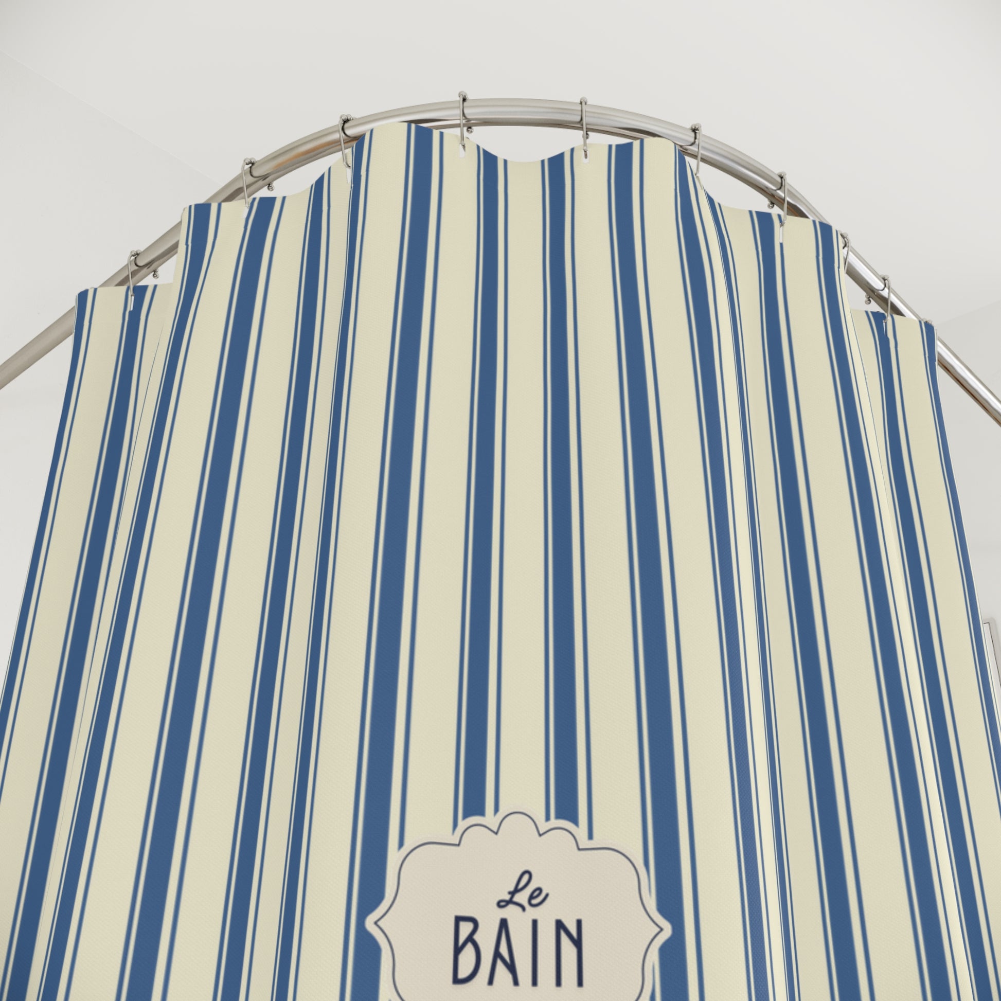 A close-up of a striped fabric shower curtain with vertical blue and cream stripes, hanging from a curved metal rod. A decorative label at the bottom reads 'Le Bain'.