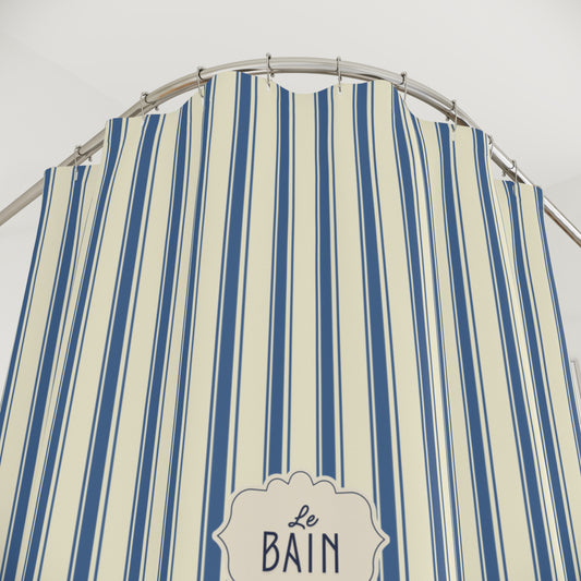 A close-up of a striped fabric shower curtain with vertical blue and cream stripes, hanging from a curved metal rod. A decorative label at the bottom reads 'Le Bain'.