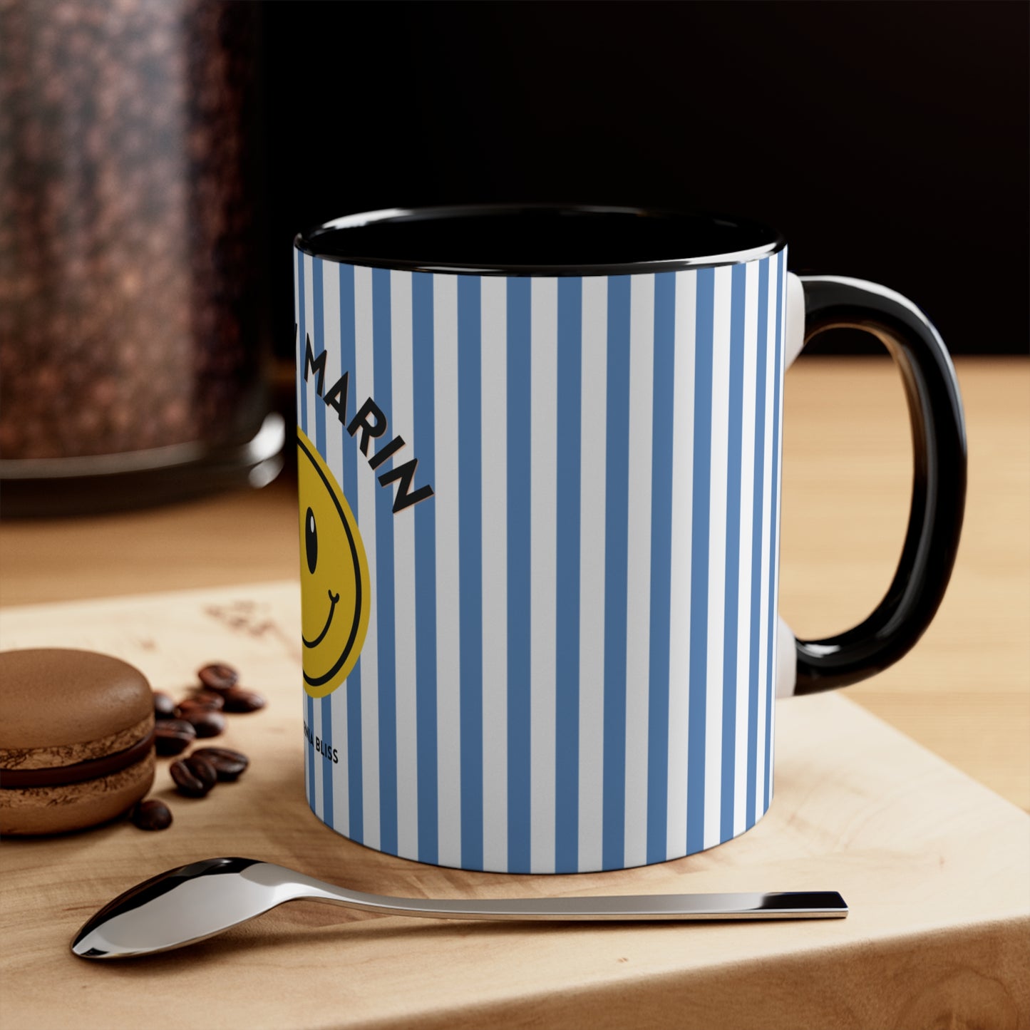 Mug with blue and white striped pattern and smiley face design on a wooden surface with coffee beans and a spoon.