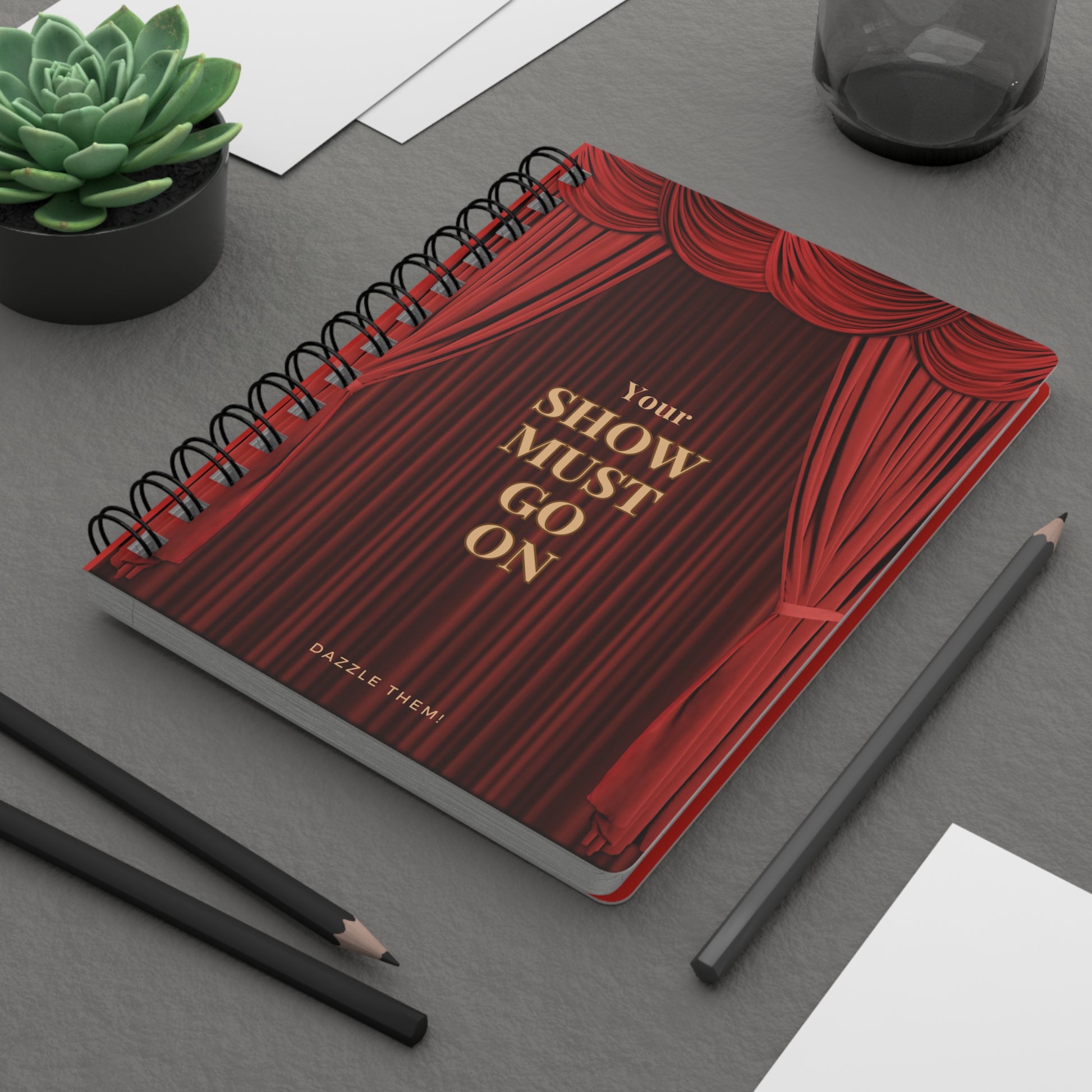 Notebook with red cover and gold text on a gray surface with pencils and a plant.