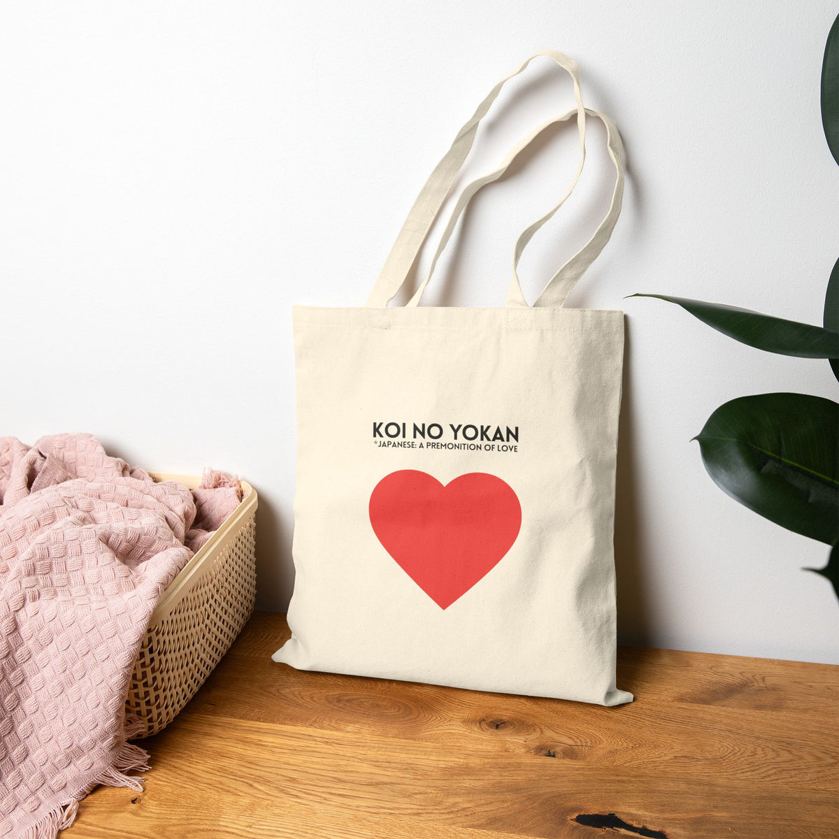 A natural canvas tote bag with long handles stands on a wooden surface against a white wall. The bag has black text reading 'KOI NO YOKAN' and a large red heart graphic.