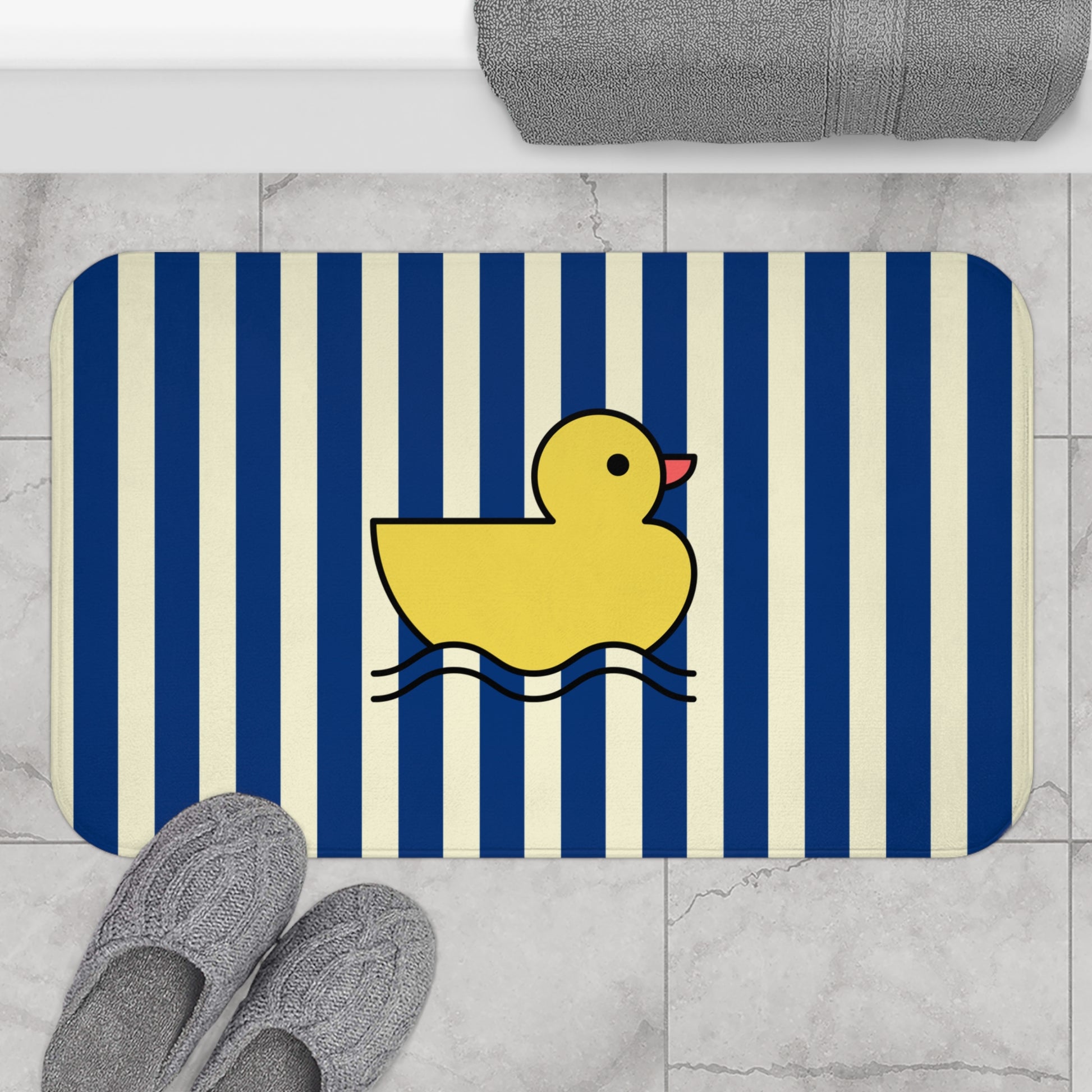 Bath mat with a yellow rubber duck on a blue and white striped background, placed on a tiled bathroom floor.