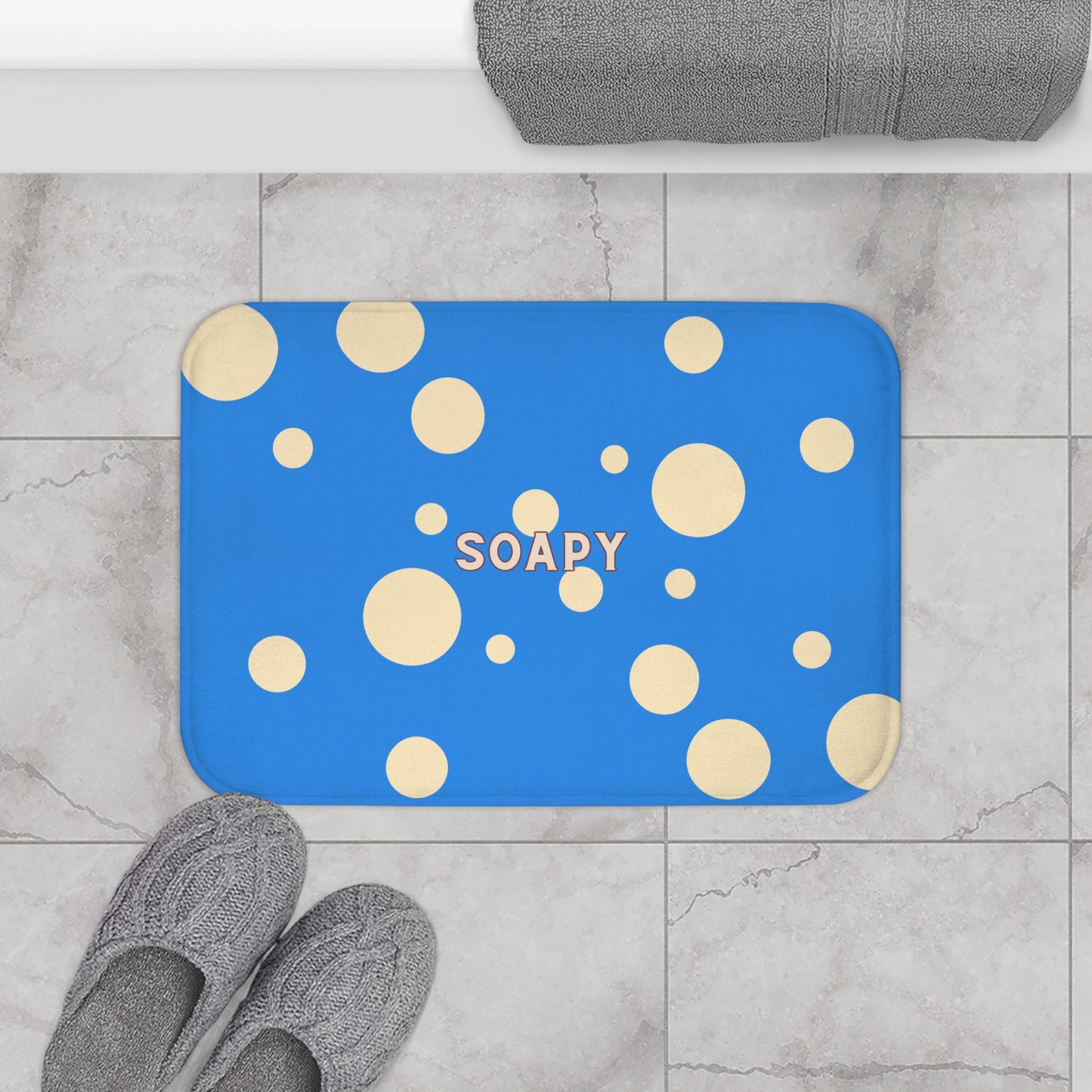 A rectangular blue bath mat with a pattern of beige and cream polka dots and the word 'SOAPY' in the center, shown on a gray marble tile floor with gray slippers and a folded gray towel nearby.