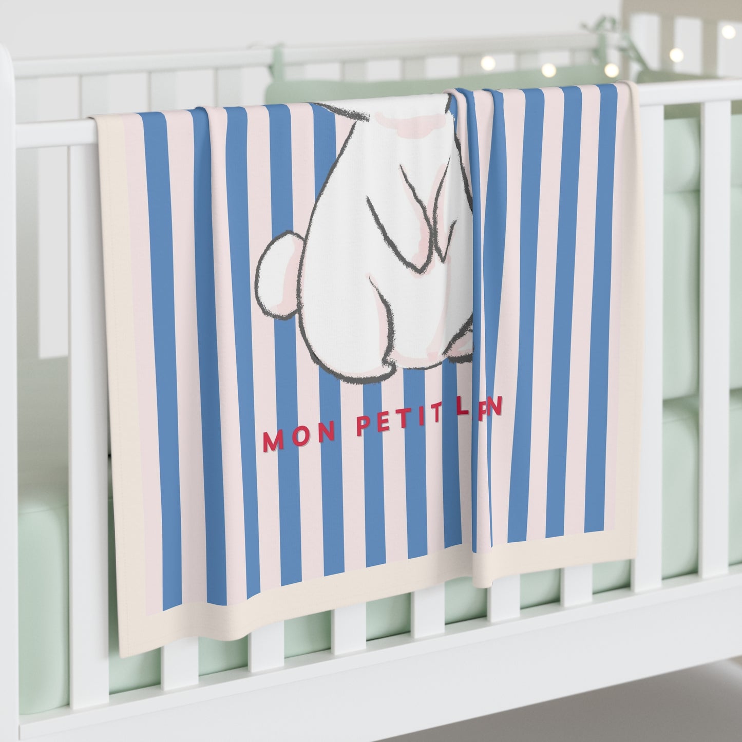 A baby blanket draped over a white crib, featuring vertical blue and cream stripes, a cartoon rabbit illustration, and the red text 'MON PETIT LUN'.