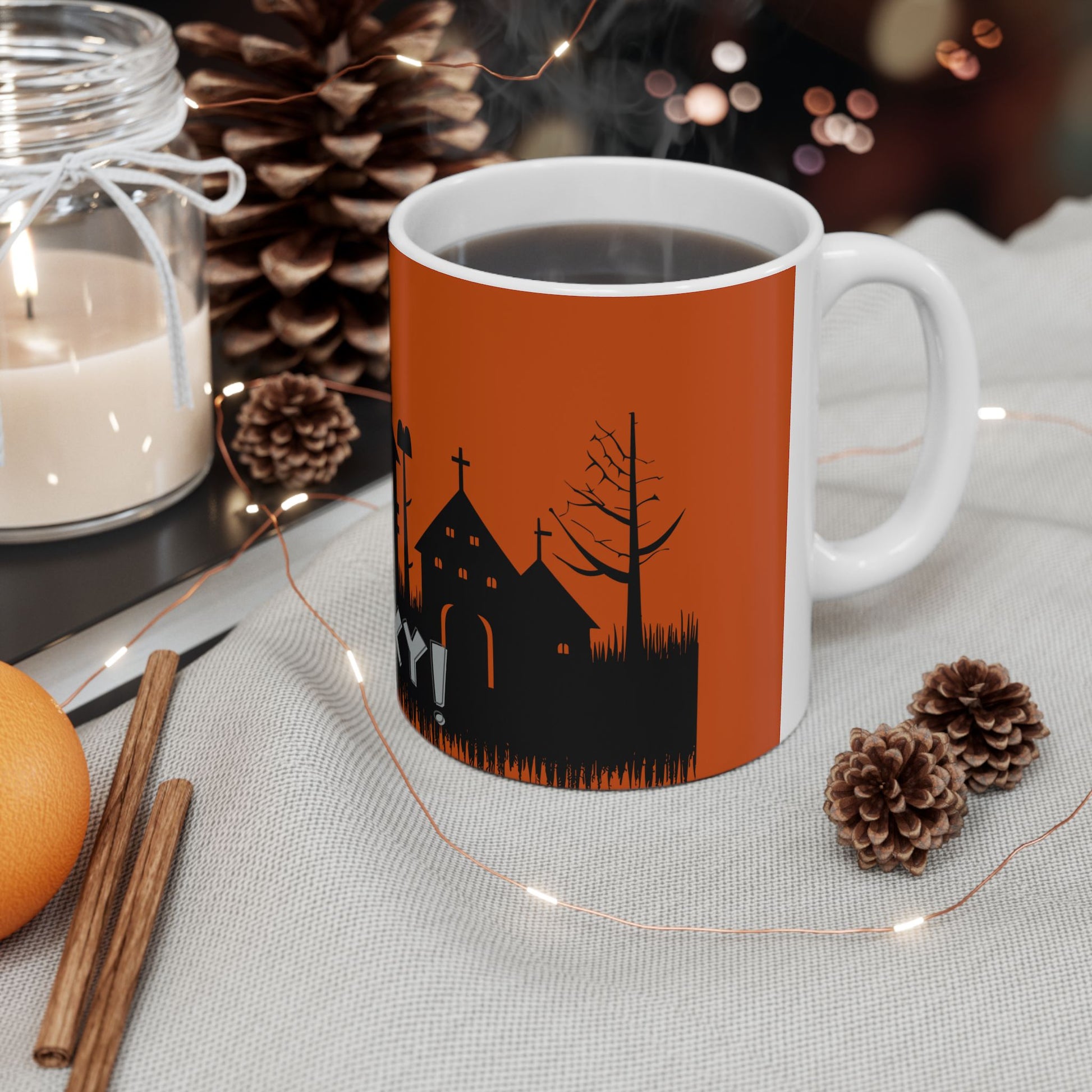 A white ceramic coffee mug with a black and orange Halloween church graphic, filled with hot coffee and set on a table with candles, pinecones, cinnamon sticks, and fairy lights.