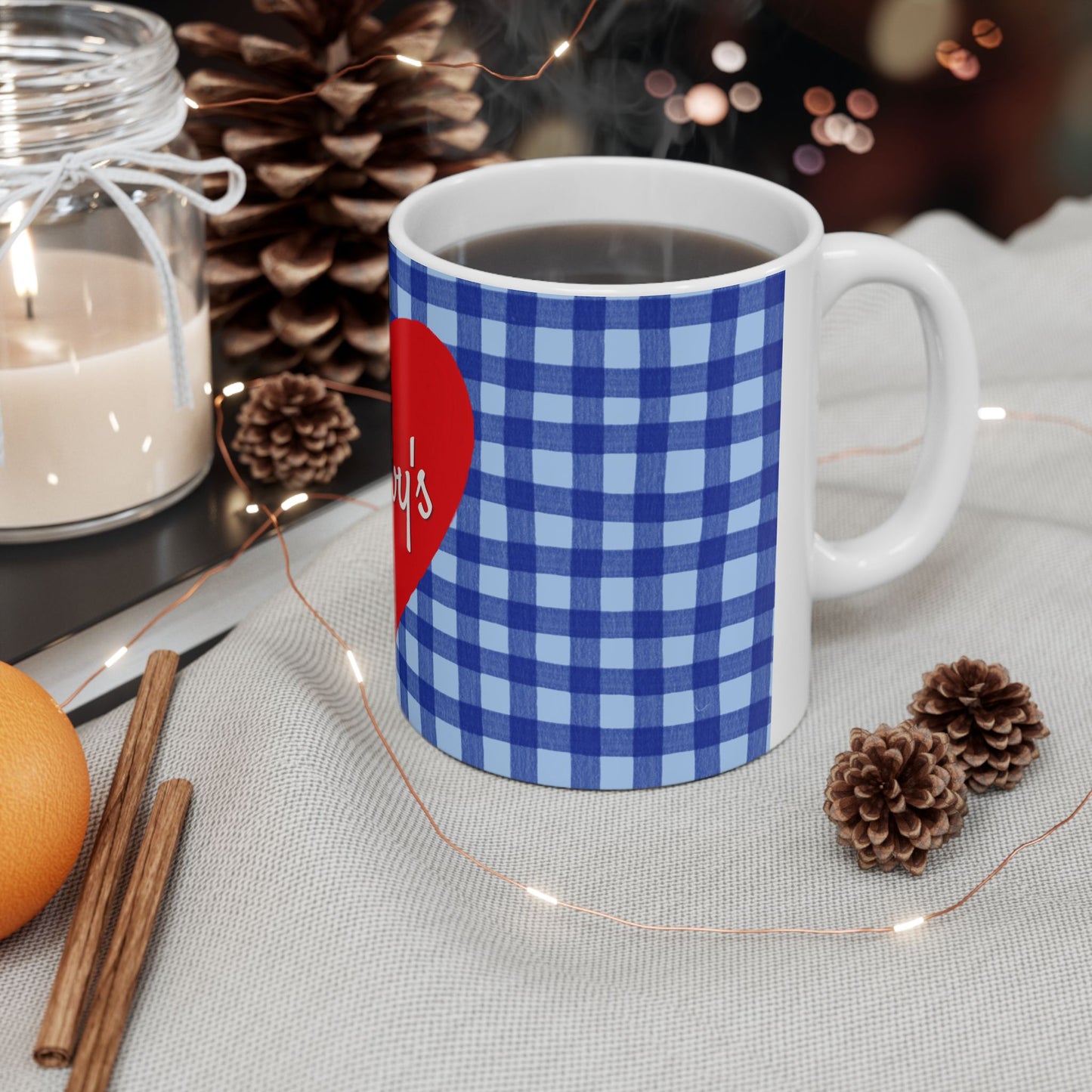 A white ceramic mug filled with a dark beverage, wrapped in a blue and light blue gingham pattern with a red heart graphic, styled on a light fabric surface with pinecones, fairy lights, a candle, cinnamon sticks, and an orange.