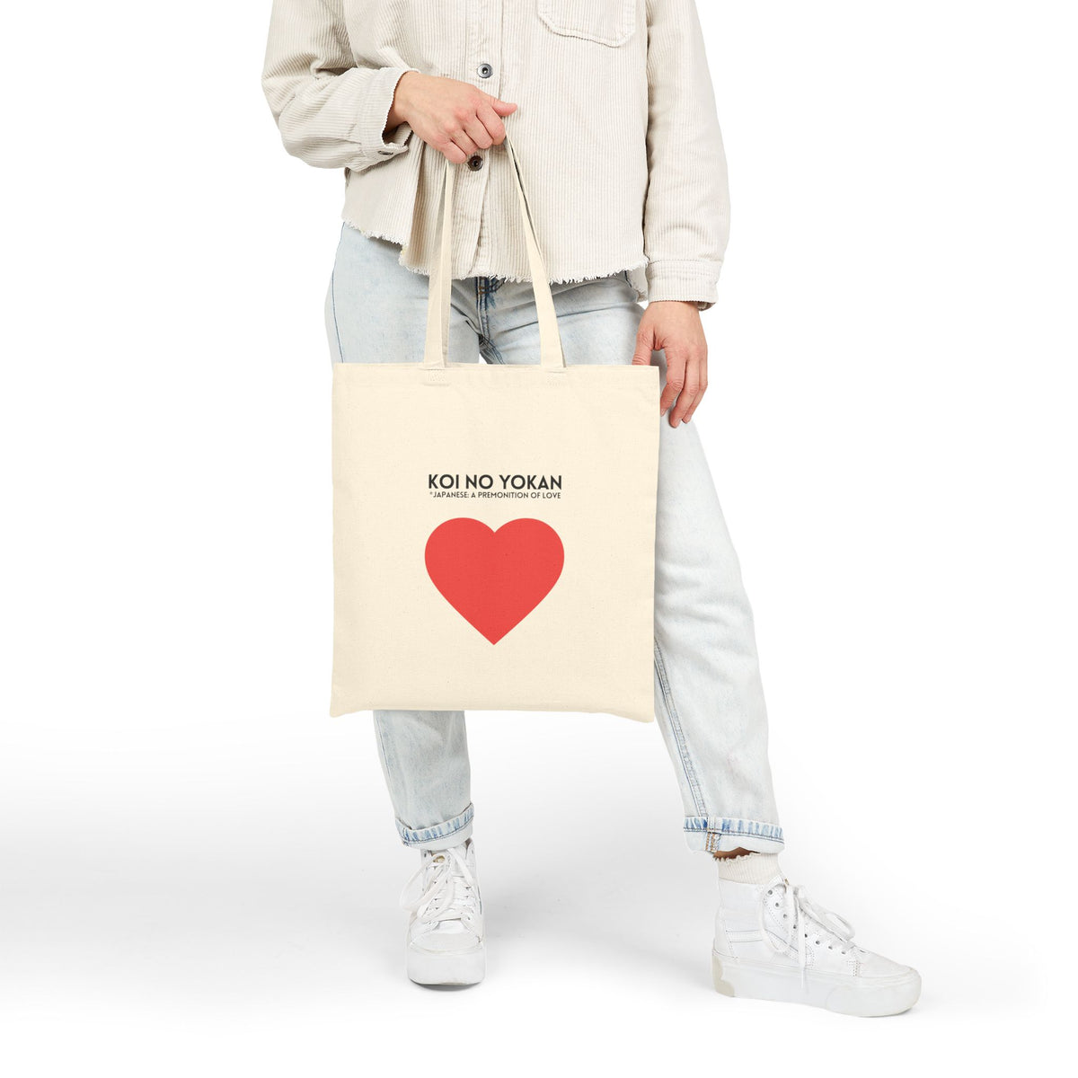 A person wearing a light beige jacket, light blue jeans, and white sneakers holds a cream canvas tote bag with a red heart graphic and the text 'KOI NO YOKAN' on a plain white studio background.