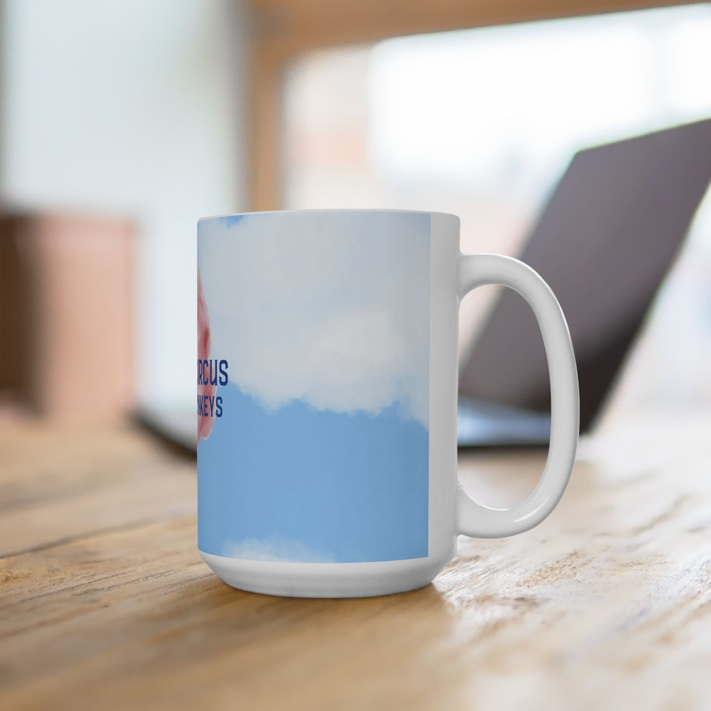 A white ceramic coffee mug with a sky-themed print featuring blue and white clouds and partial red text, sitting on a wooden table with a blurred laptop in the background.