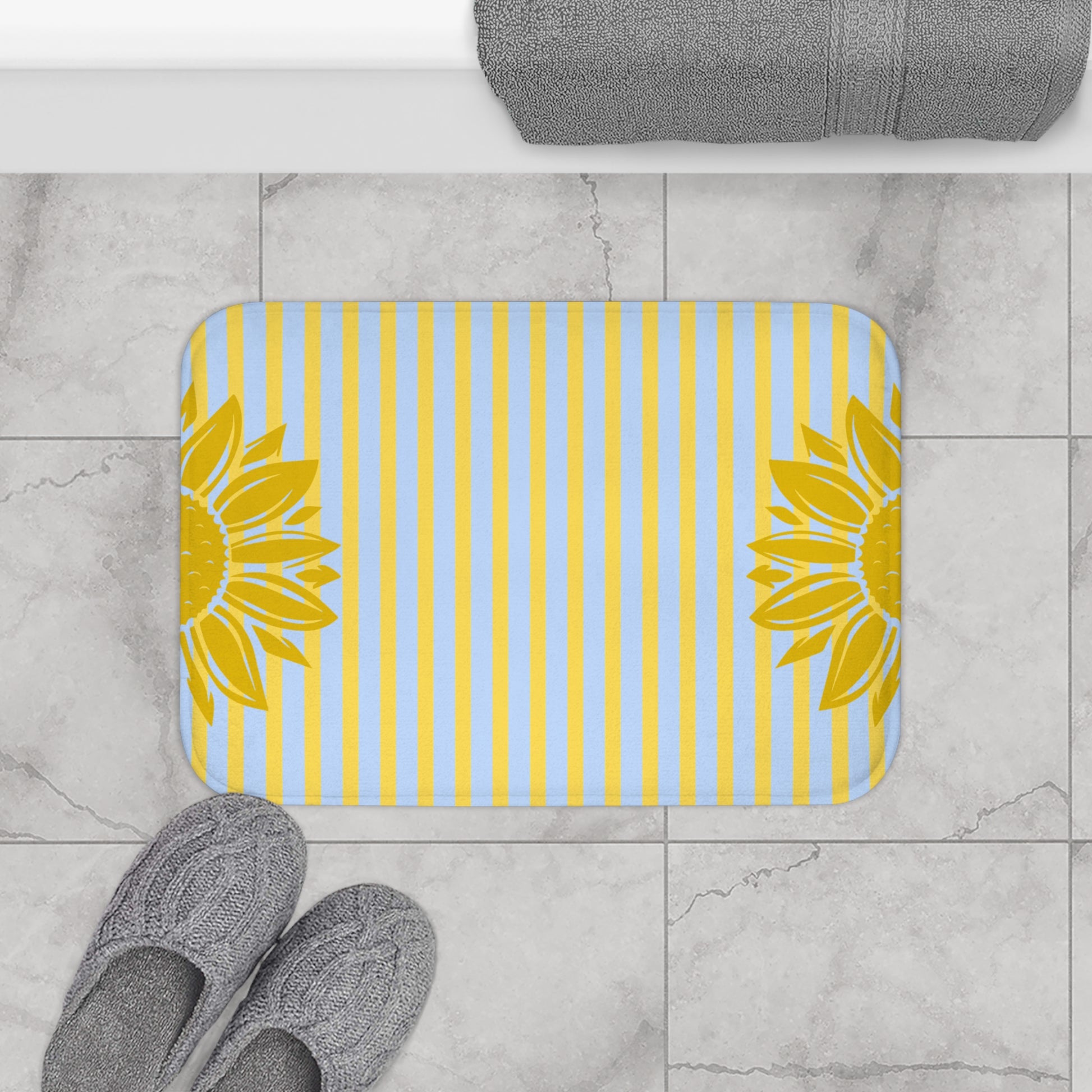 Bath mat with sunflower design on a striped pattern, placed on a tiled floor with slippers and a towel.