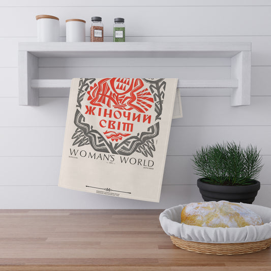 A beige kitchen towel with a vintage-style red and black graphic reading 'Woman's World' hangs from a white wall-mounted rack in a bright kitchen. A small potted green plant and a woven basket with a loaf of bread sit on the wooden floor below.