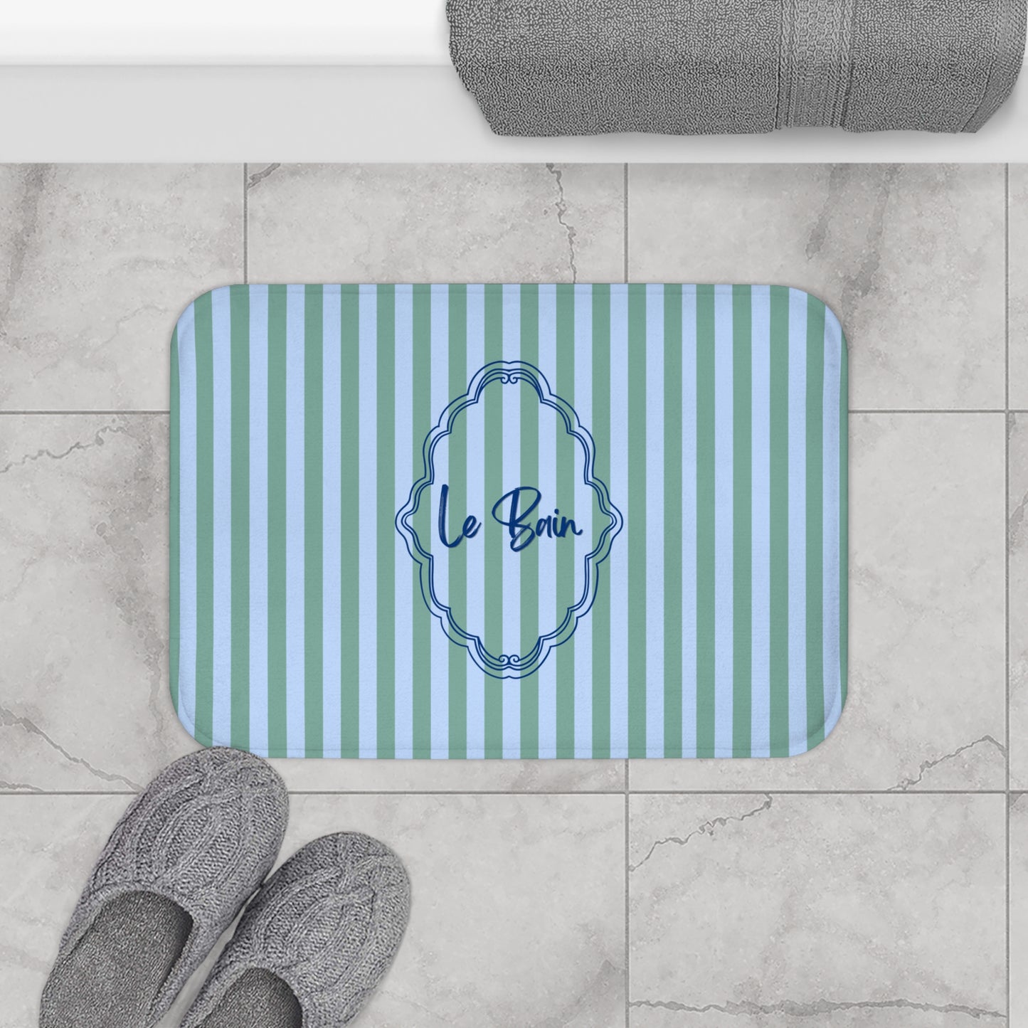 Striped bath mat with 'Le Bain' text on a tiled floor with slippers and a towel.