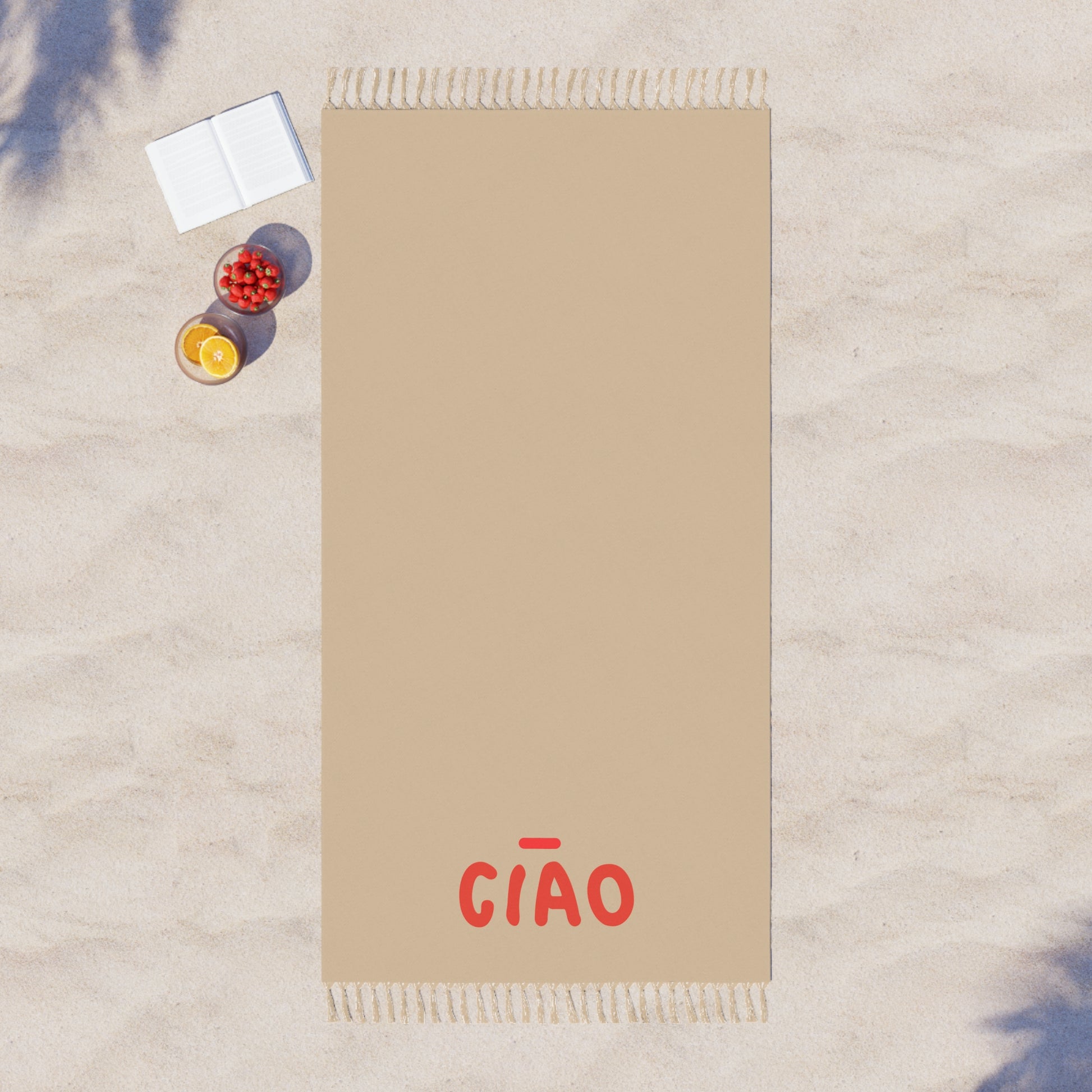 Beige towel with 'CIAO' text on a sandy surface with a book and fruit.