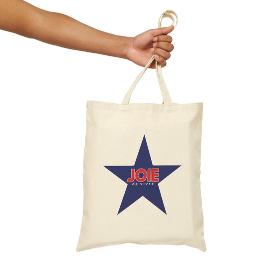 An off-white canvas tote bag with a large navy blue star graphic on the front, featuring the word 'JOIE' in red and 'de vivre' in white. A person's arm and hand are holding the bag by its top handle against a plain white background.