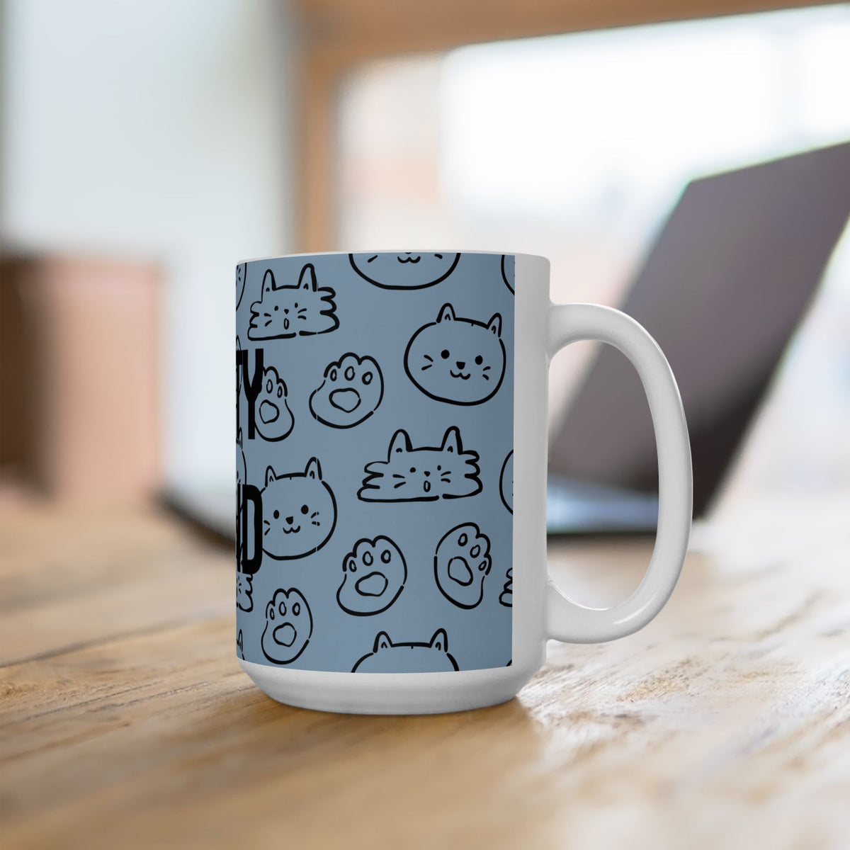 A white ceramic mug with a light blue printed wrap featuring black cartoon cat faces and paw prints, sitting on a wooden table with a blurred laptop in the background.