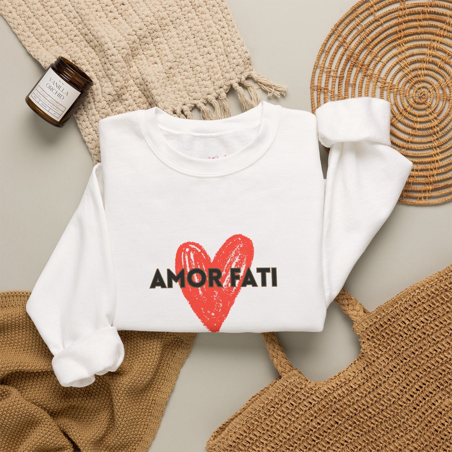 A folded white long-sleeve sweatshirt with a red heart graphic and the text 'AMOR FATI' printed across the chest, styled in a flat lay with a woven blanket, a small candle, and a woven mat on a light neutral background.