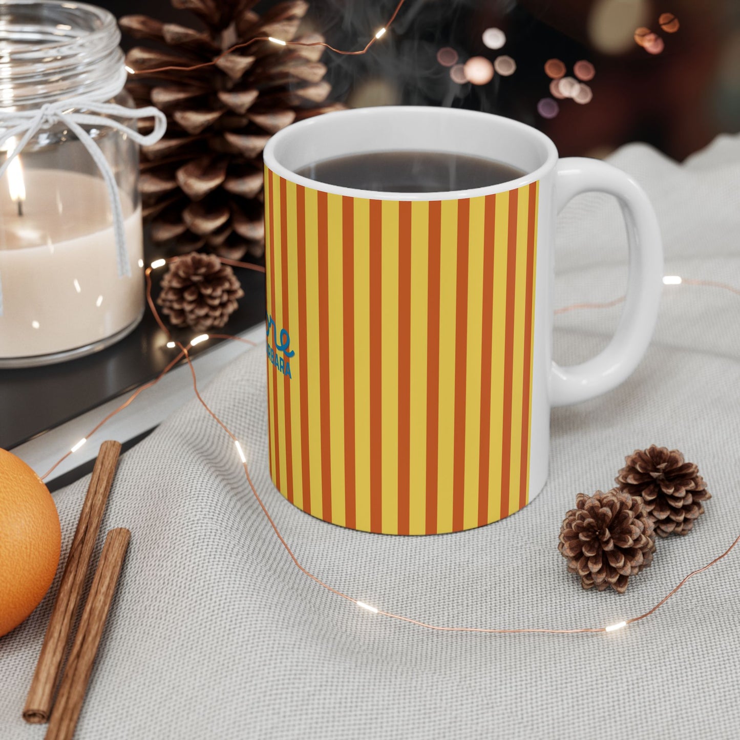 A white ceramic mug with yellow and orange vertical stripes sits on a light gray fabric surface, surrounded by cozy props including pinecones, a lit candle in a glass jar, cinnamon sticks, and warm fairy lights.