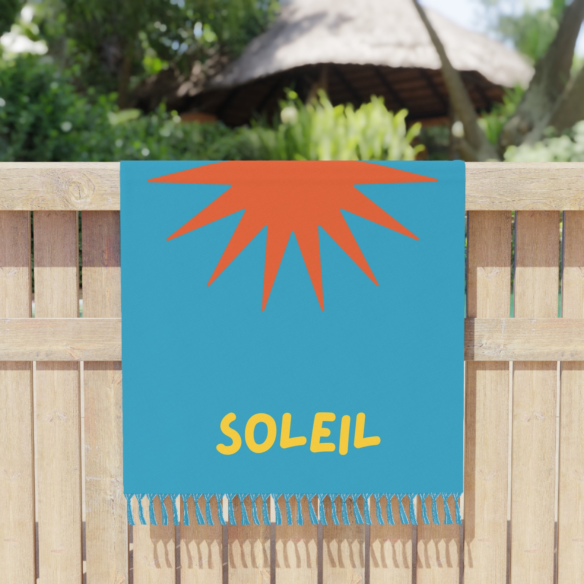 Beach towel with orange sun design and 'Soleil' text on a blue background, hanging on a wooden fence.