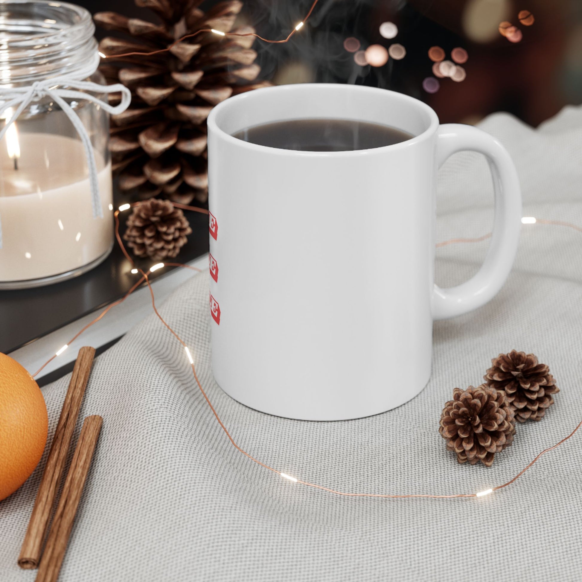 White mug with red text on a table with decorative items including a candle, oranges, and pine cones.