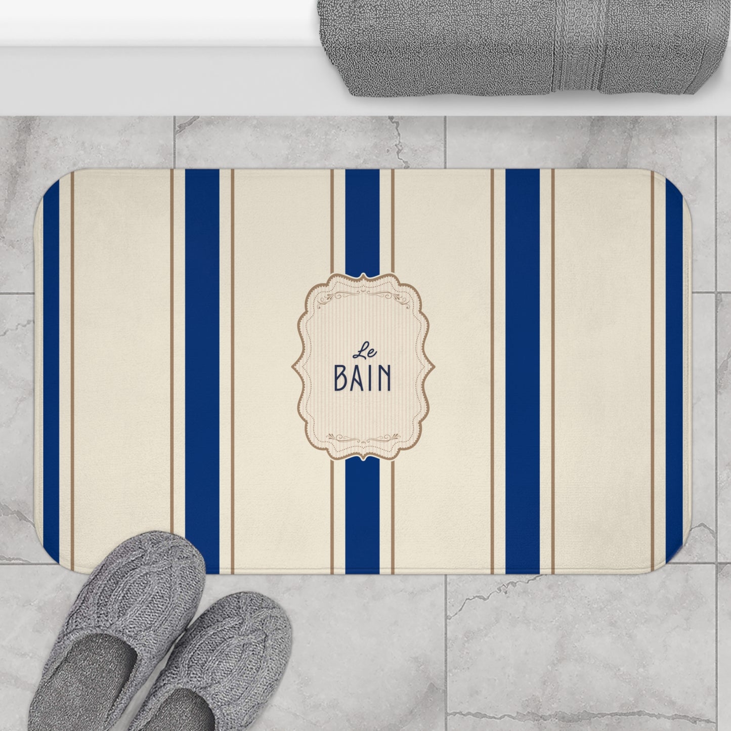 Bath mat with blue stripes and 'Le Bain' text on a marble floor with slippers and a towel.