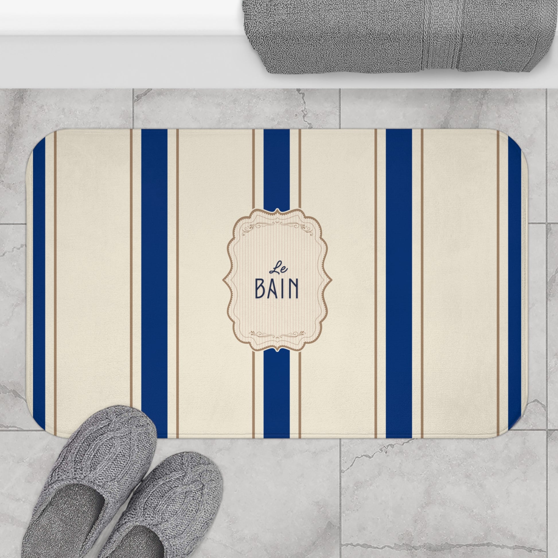 Bath mat with blue stripes and 'Le Bain' text on a marble floor with slippers and a towel.