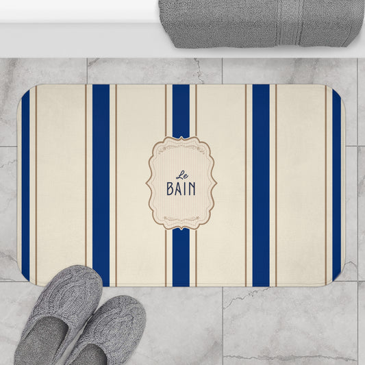Bath mat with blue stripes and 'Le Bain' text on a marble floor with slippers and a towel.