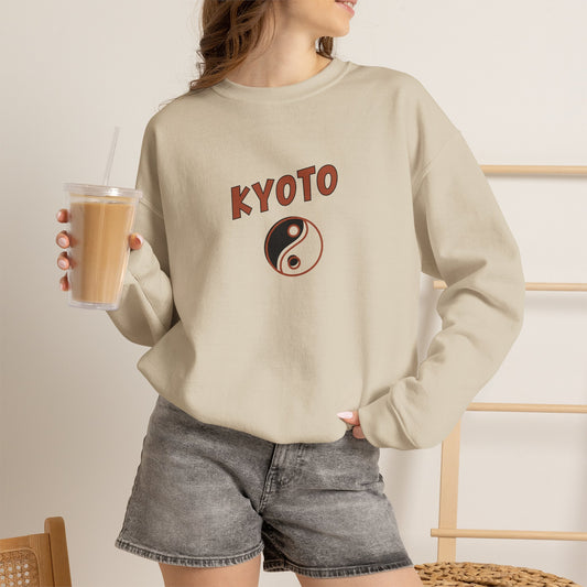 Person wearing a beige sweatshirt with 'KYOTO' and a yin yang symbol, holding a drink.