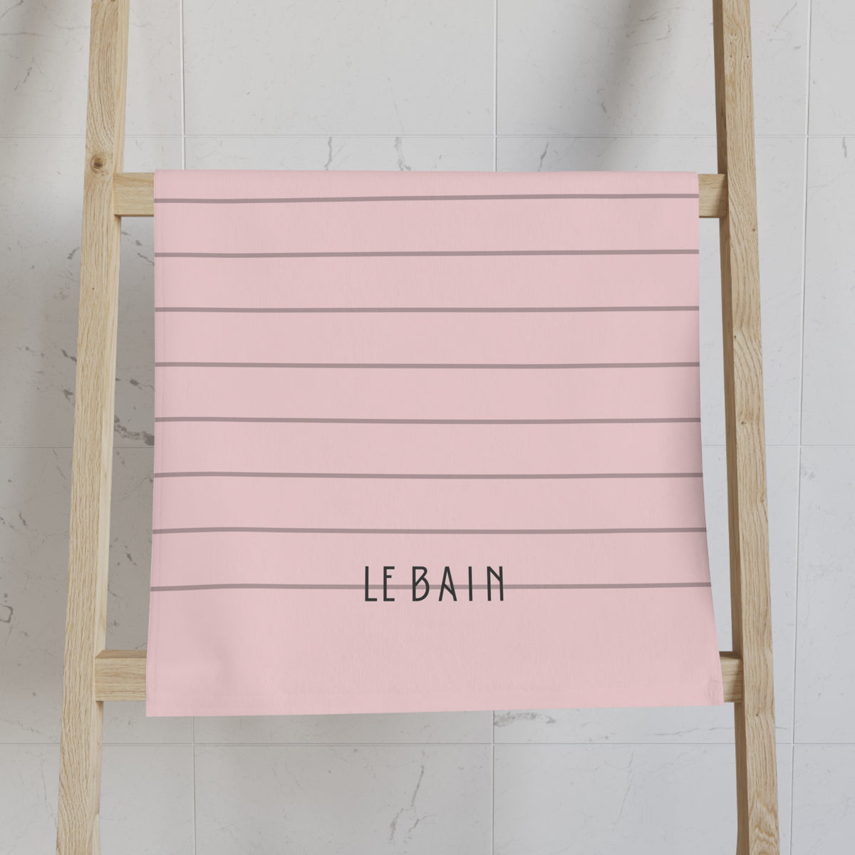 A light pink towel with thin dark horizontal stripes and the word 'LE BAIN' printed in black at the bottom, hanging on a wooden ladder against a white tiled wall.
