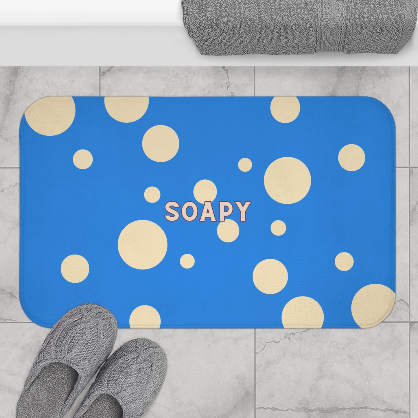 A rectangular blue bath mat with rounded corners, covered in scattered cream polka dots and the word “SOAPY” in the center, shown on a light marble floor with gray slippers and a folded gray towel nearby.