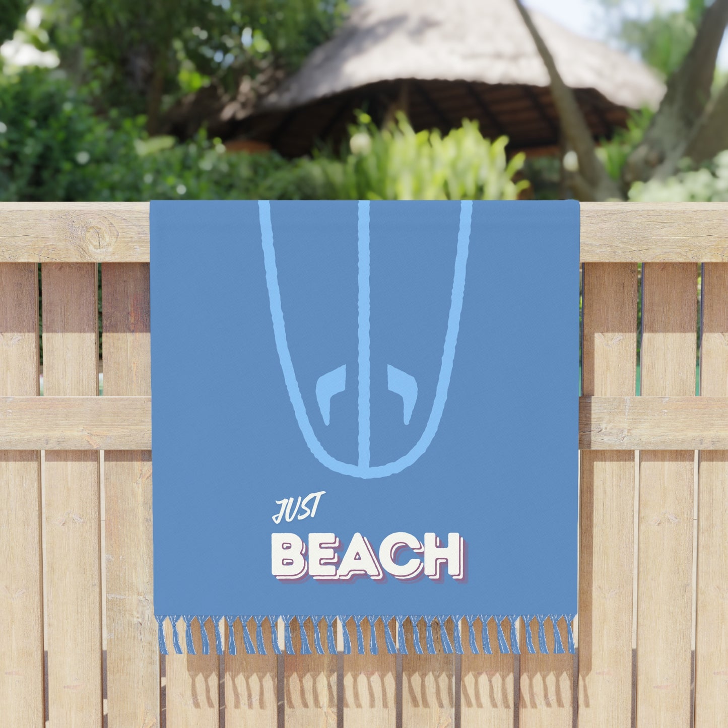 Blue beach towel with 'Just Beach' text and surfboard design on a wooden fence background.