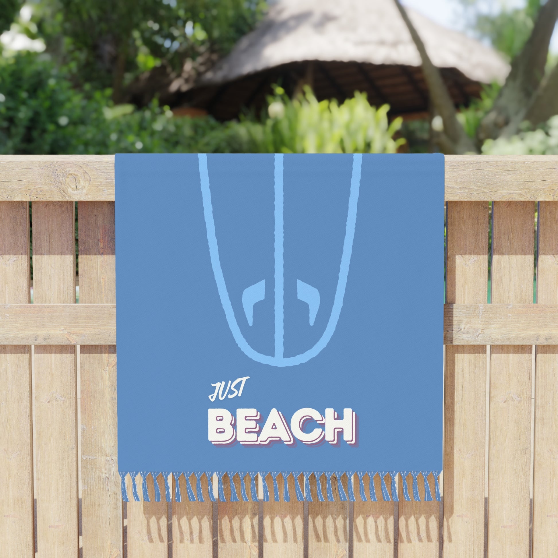 Blue beach towel with 'Just Beach' text and surfboard design on a wooden fence background.
