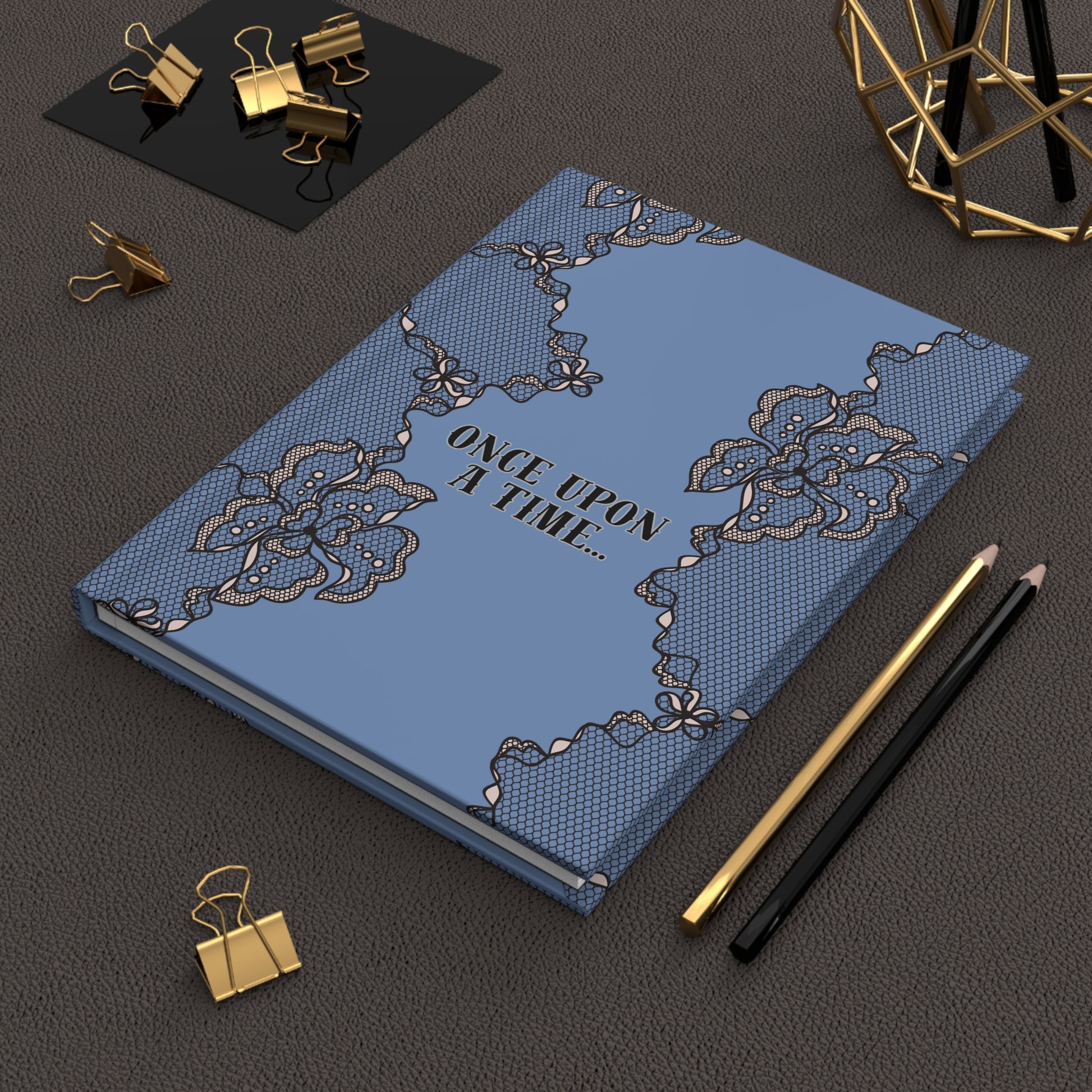 Blue notebook with lace design and 'Once Upon a Time' text on a dark surface with gold clips and pencils.