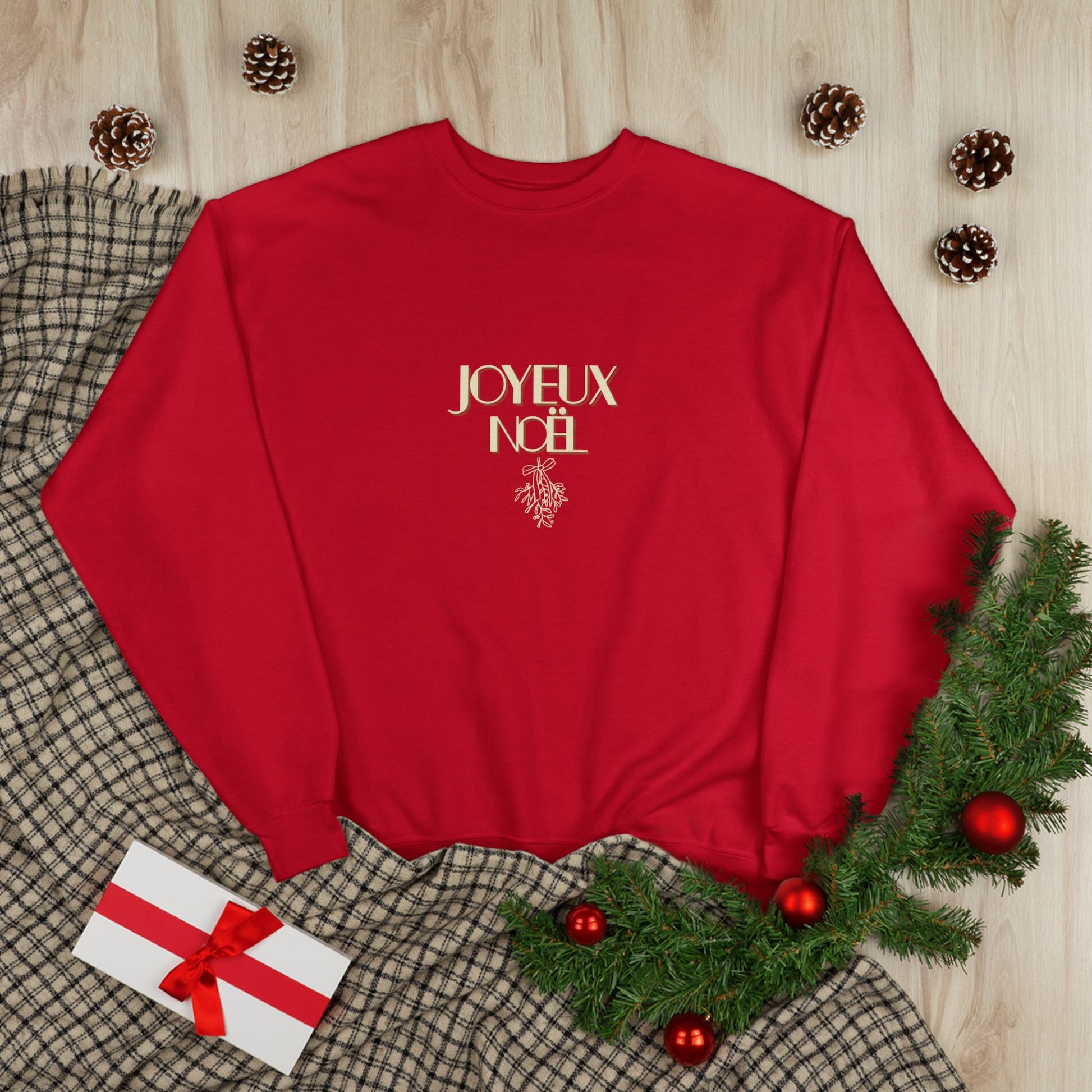 Red sweatshirt with 'JOYEUX NOEL' text on a wooden surface with Christmas decorations.