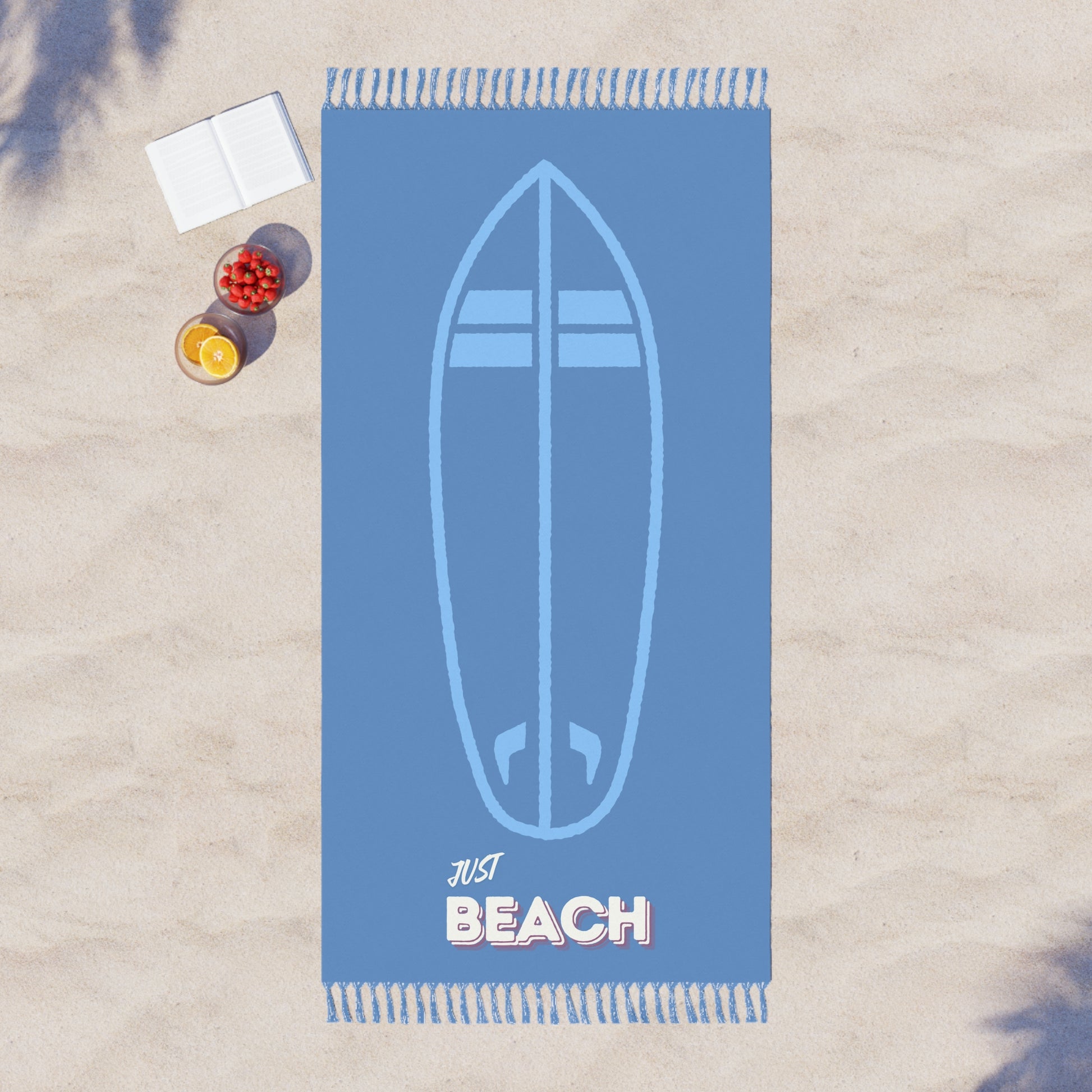 Blue beach towel with surfboard design and 'Just Beach' text on a sandy background