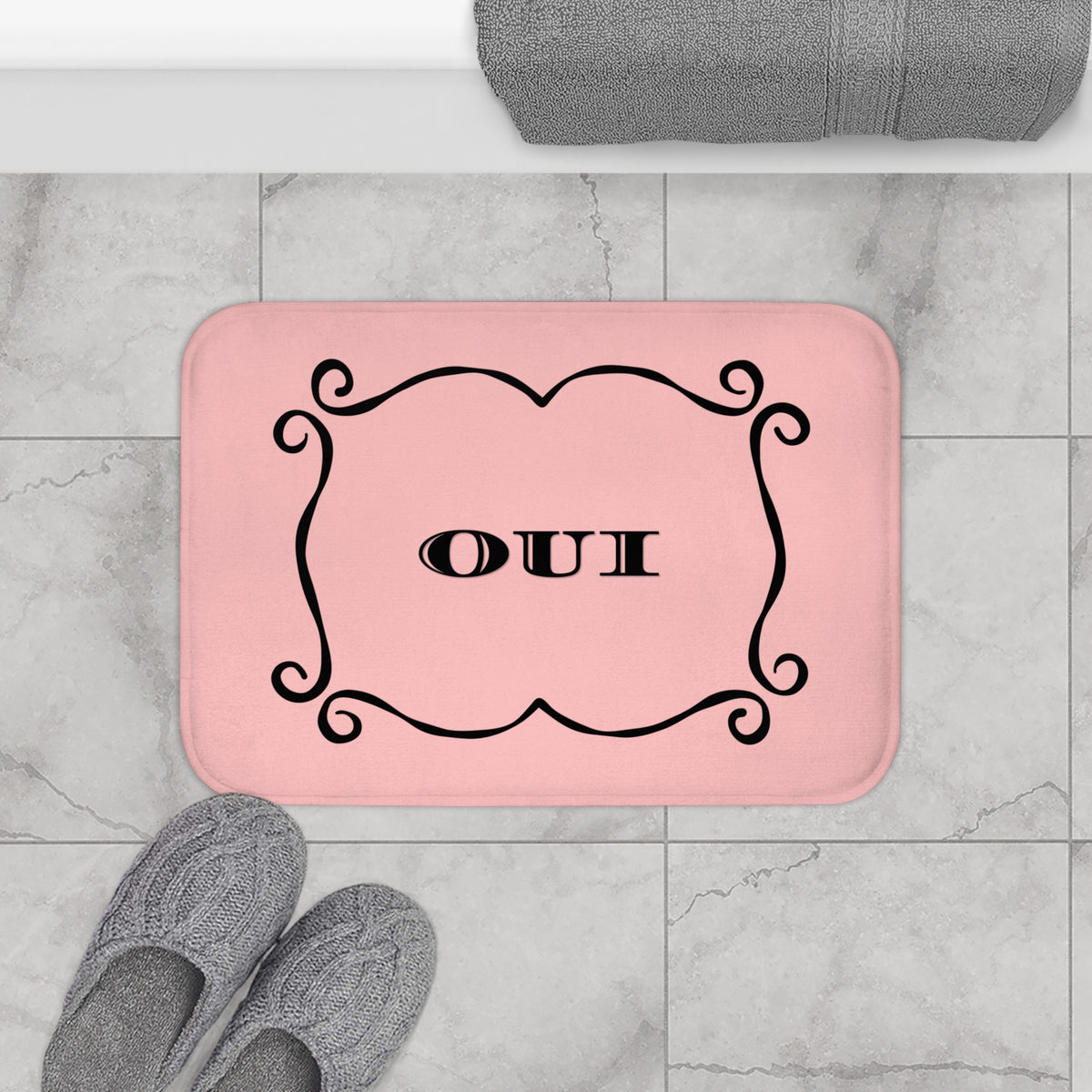 A pink rectangular bath mat with rounded corners sits on a light gray marble tile floor. The mat has a black ornate frame and the word 'OUI' in the center.