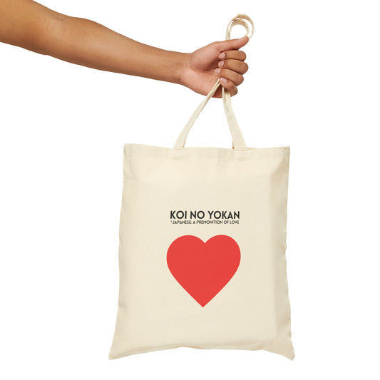 An off-white canvas tote bag with a red heart and black text reading 'KOI NO YOKAN -JAPANESE A PROPHETION OF LOVE-' held by a person's hand against a solid white background.