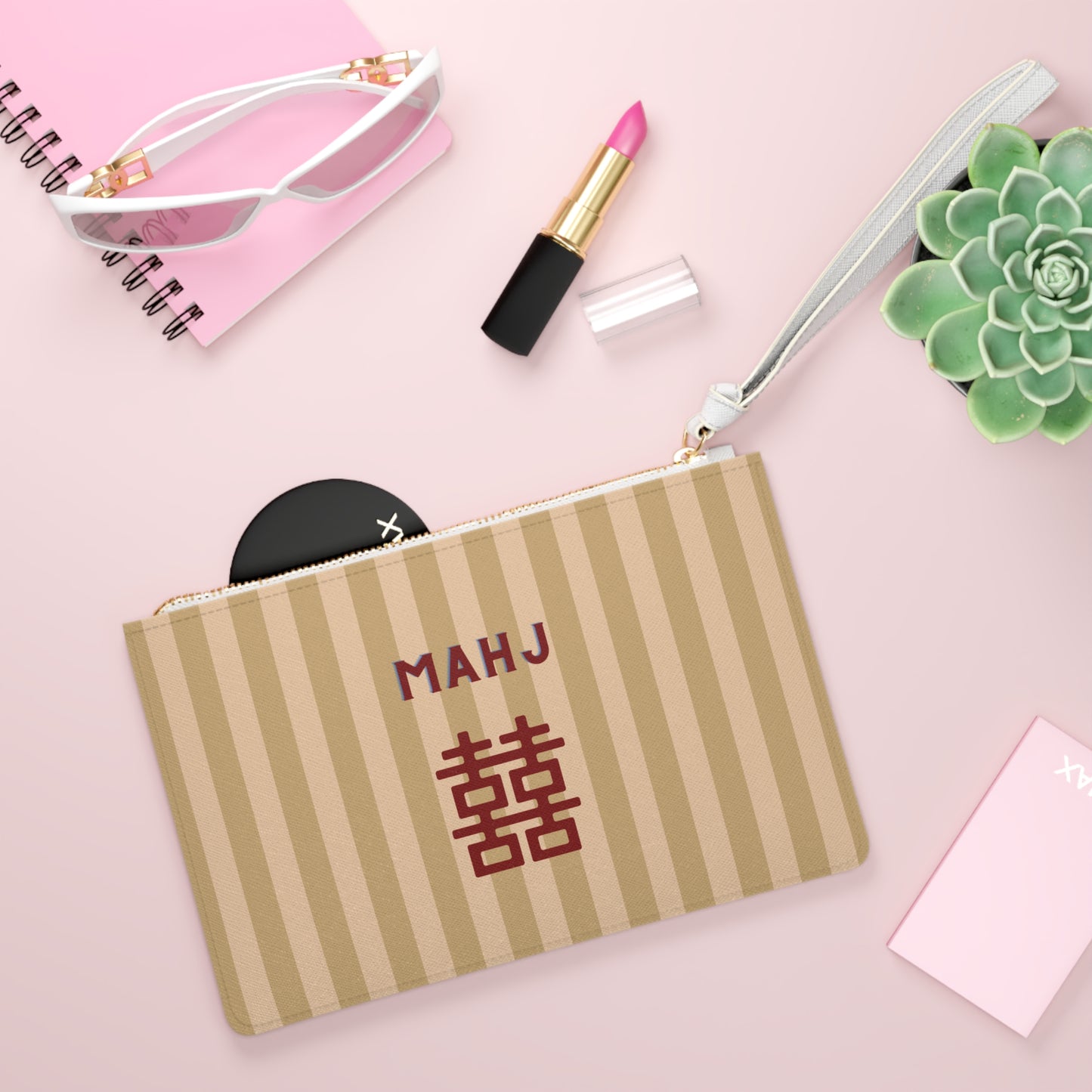 A personalized zippered pouch with vertical beige and brown stripes, featuring the text 'MAHJ' and a Chinese double happiness symbol in dark red. The pouch is styled in a flat-lay arrangement on a pink background alongside sunglasses, lipstick, a succulent, and a notebook.