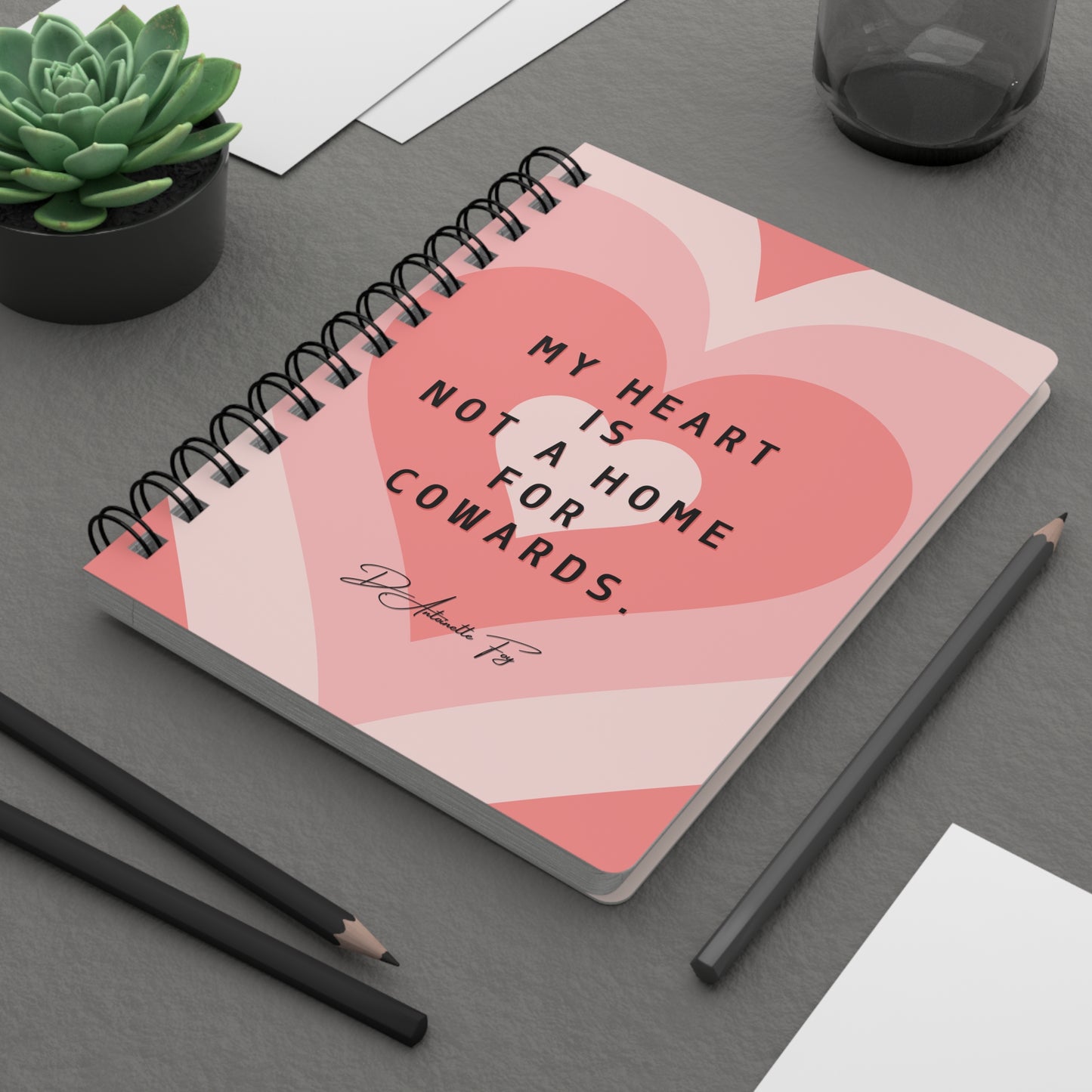 Notebook with heart design and quote on a gray surface with pencils and a plant.