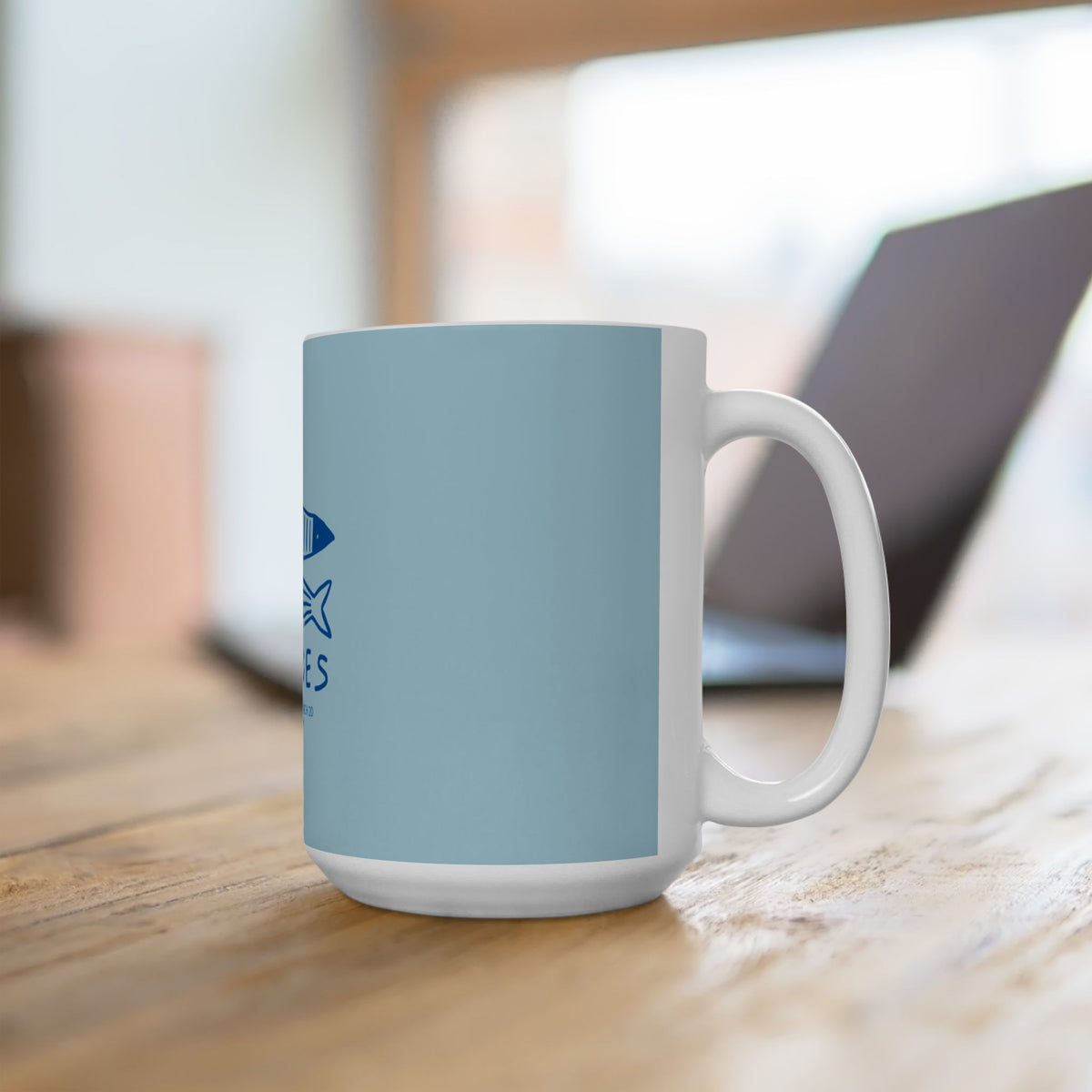 A white ceramic coffee mug with a light blue wrap-around design, featuring a dark blue shark graphic and the text 'SHARKS' on the side, sitting on a wooden table with a blurred laptop in the background.