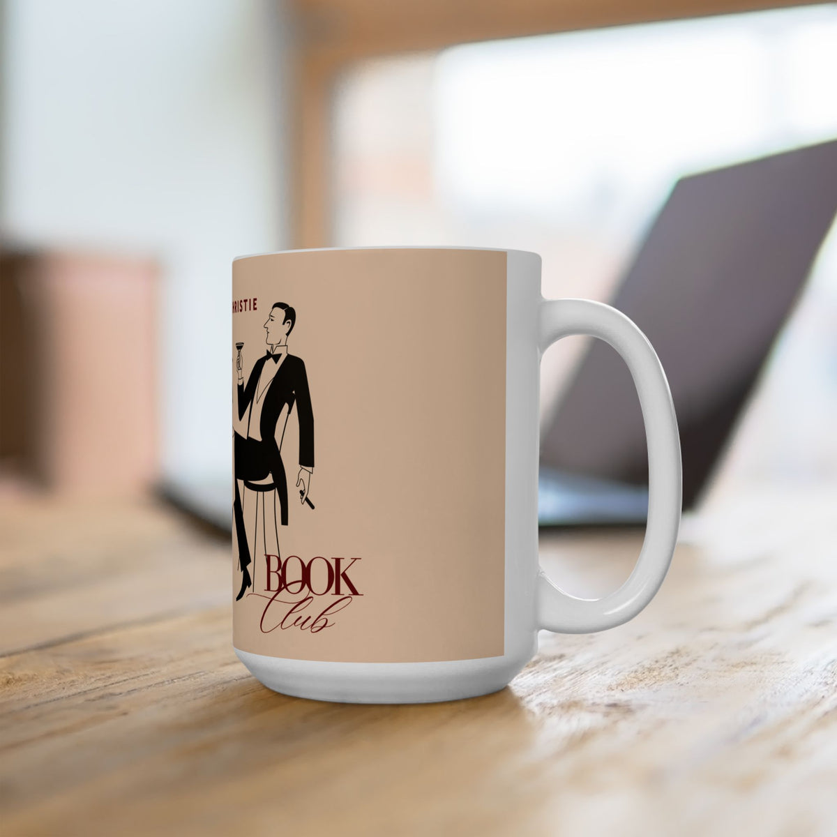 A white ceramic mug with a beige graphic print featuring a tuxedo-wearing figure and the words 'The Book Club,' placed on a wooden table with a blurred laptop in the background.