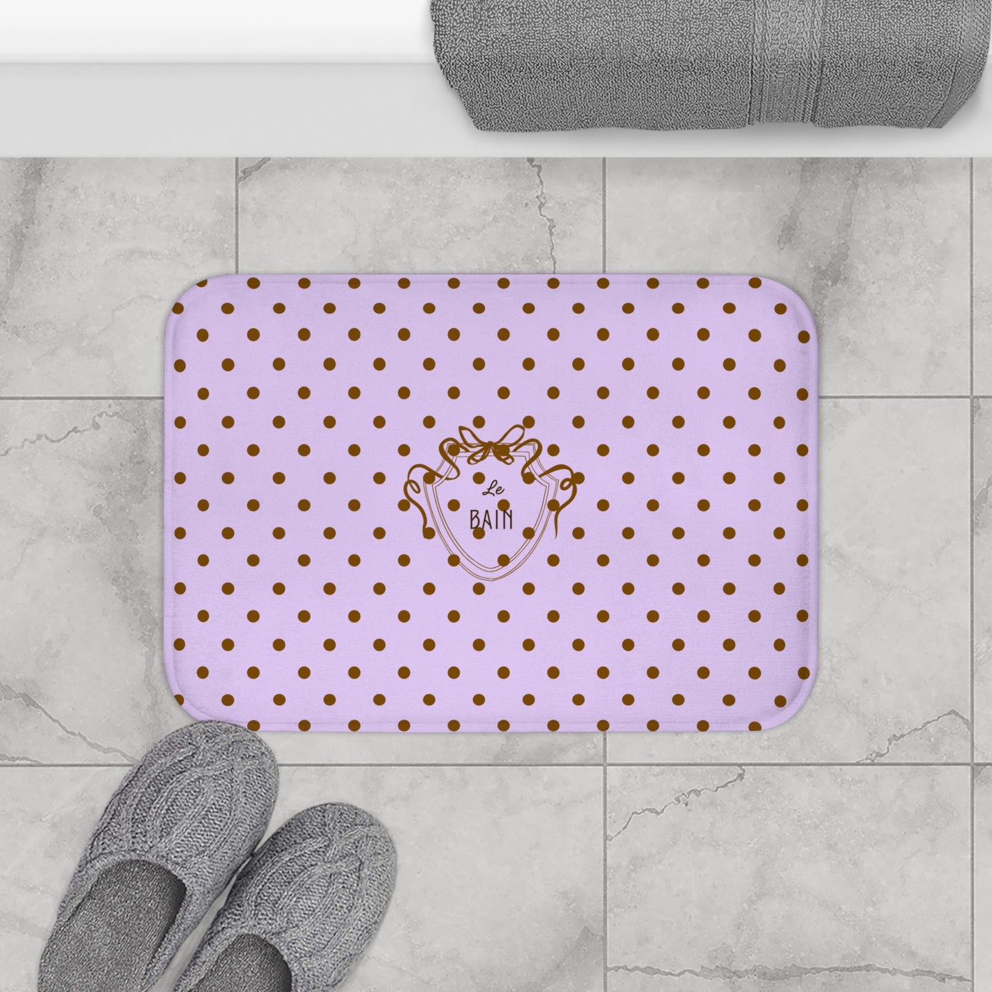 Purple bath mat with gold polka dots and a logo on a marble floor with gray slippers and a towel.