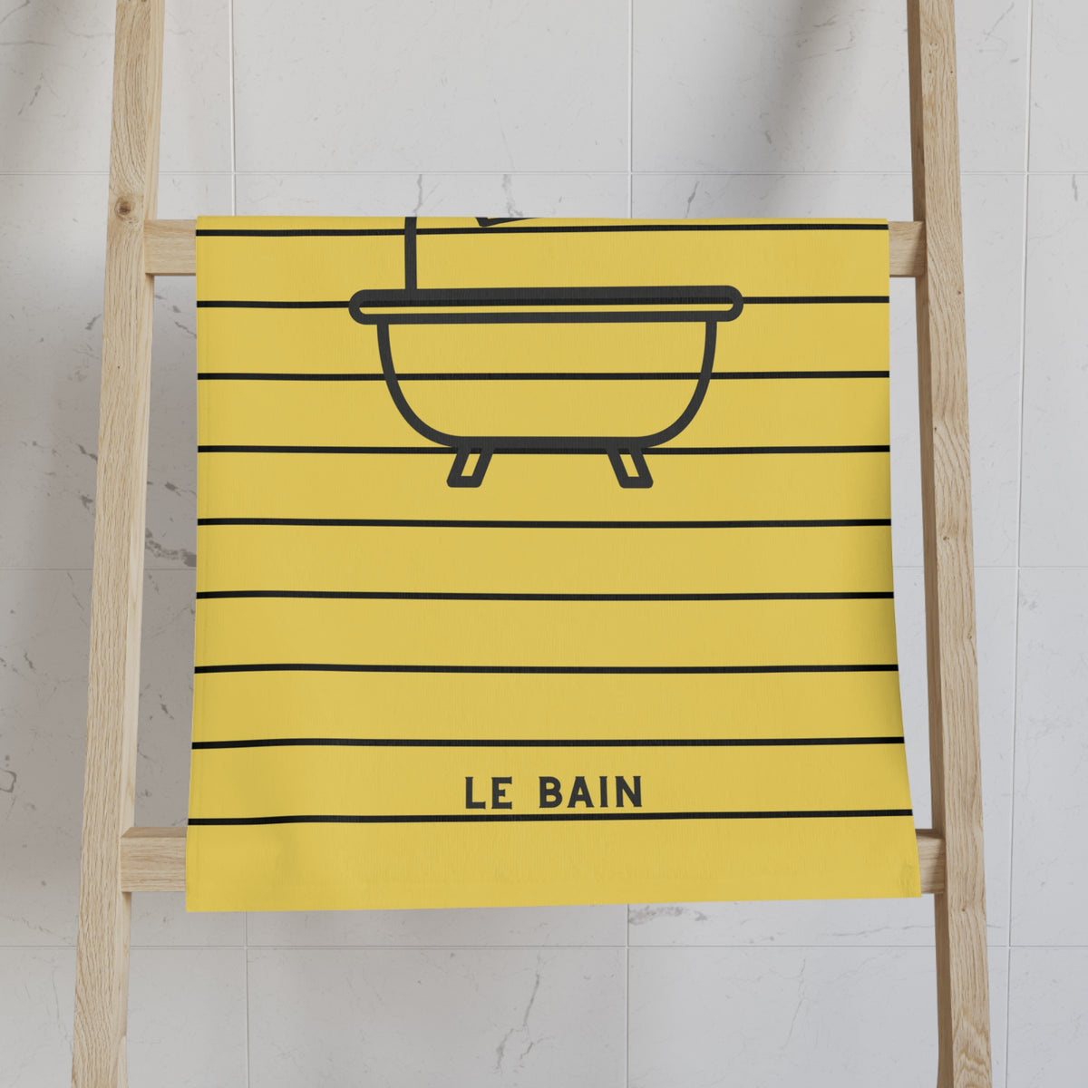 A yellow kitchen towel with a black horizontal stripe pattern and a bathtub outline, featuring the text 'LE BAIN' at the bottom, hanging on a wooden ladder against a white tiled wall.