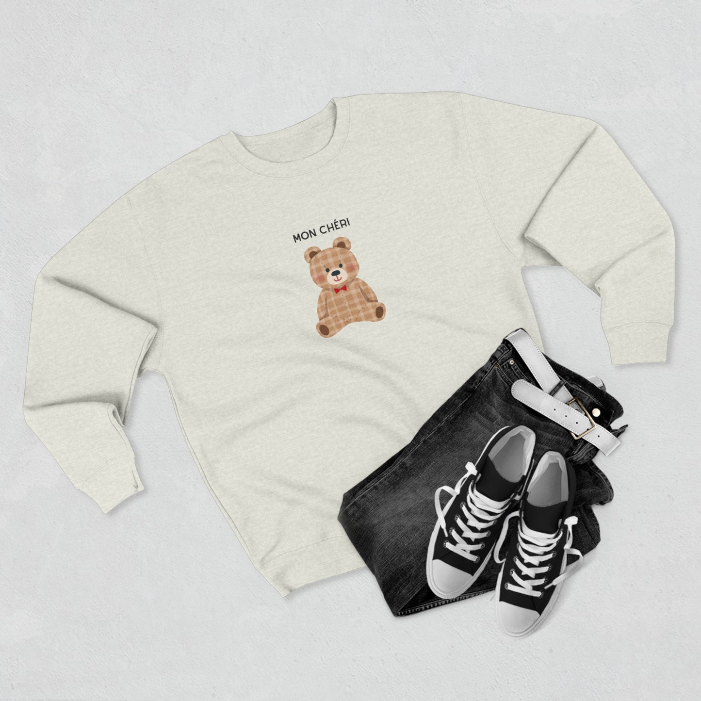 White sweatshirt with teddy bear design, black jeans, and black sneakers on a light gray background