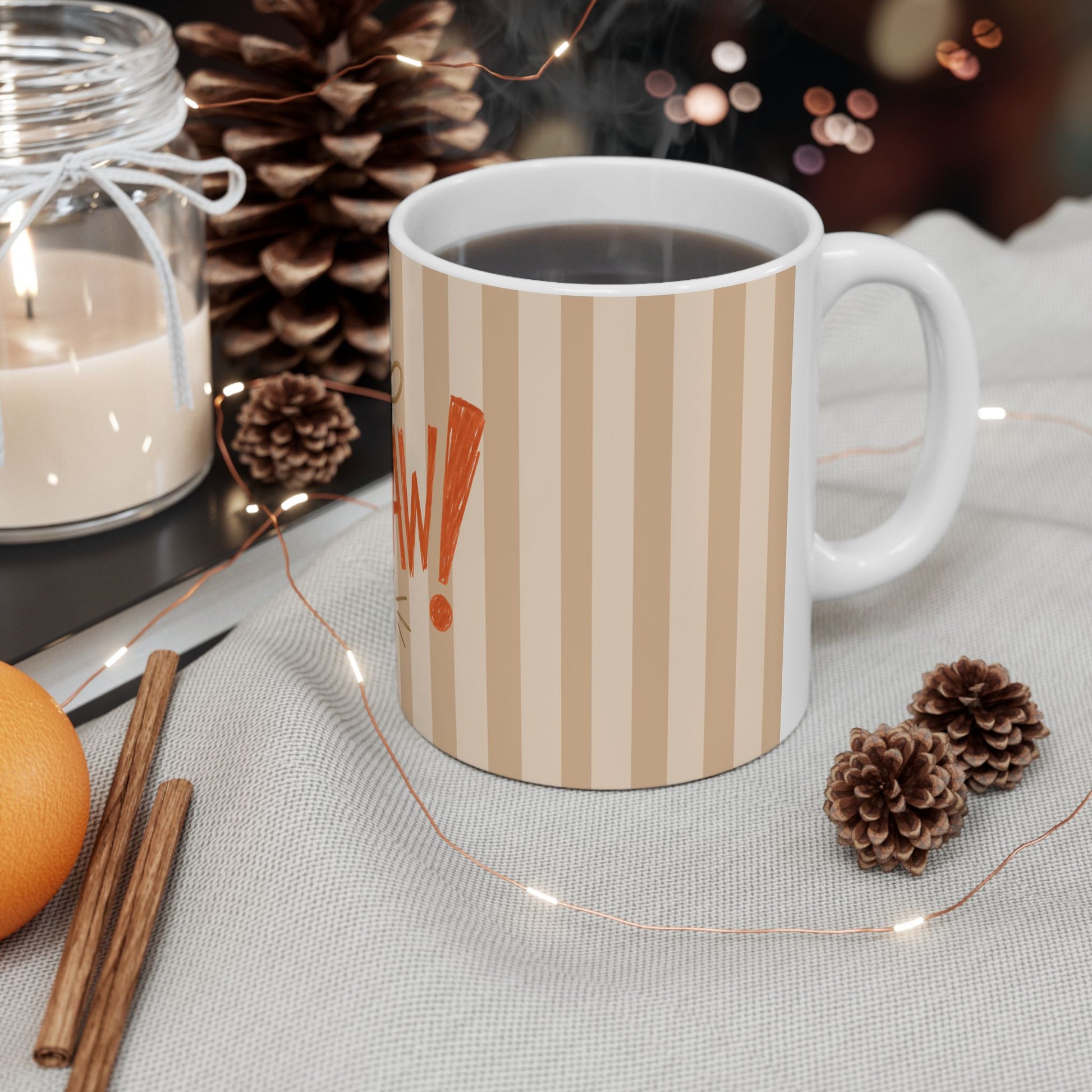 A white ceramic mug with beige vertical stripes and an orange exclamation mark graphic, filled with a dark beverage and set on a light fabric surface with pinecones, cinnamon sticks, a candle jar, and warm string lights.