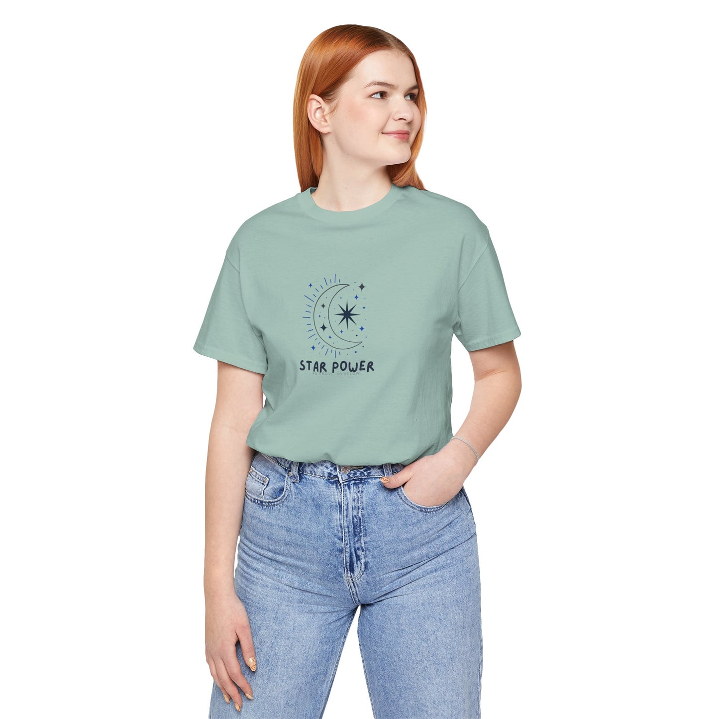A woman with red hair wearing a sage green graphic t-shirt with a crescent moon and star design, paired with light blue jeans, against a plain white background.