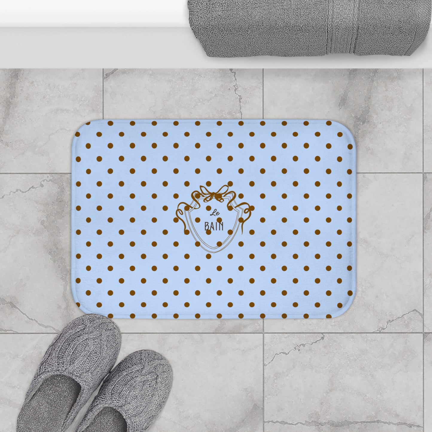 Blue bath mat with gold polka dots and a logo on a tiled bathroom floor.